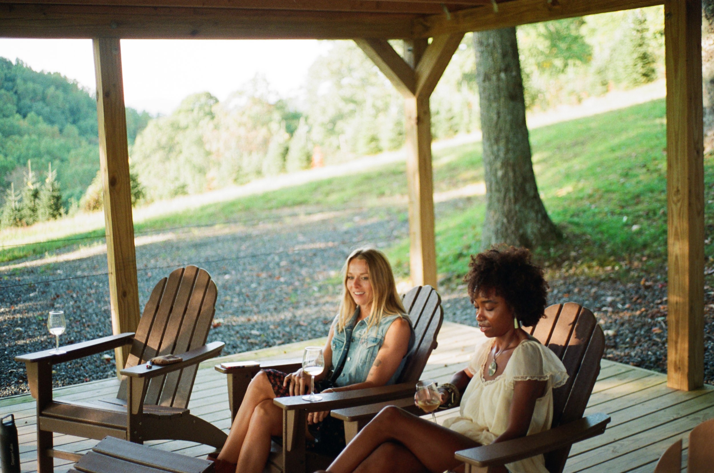 Two women sitting on Adirondack chairs on a wooden porch, holding wine glasses, with a scenic green landscape in the background.