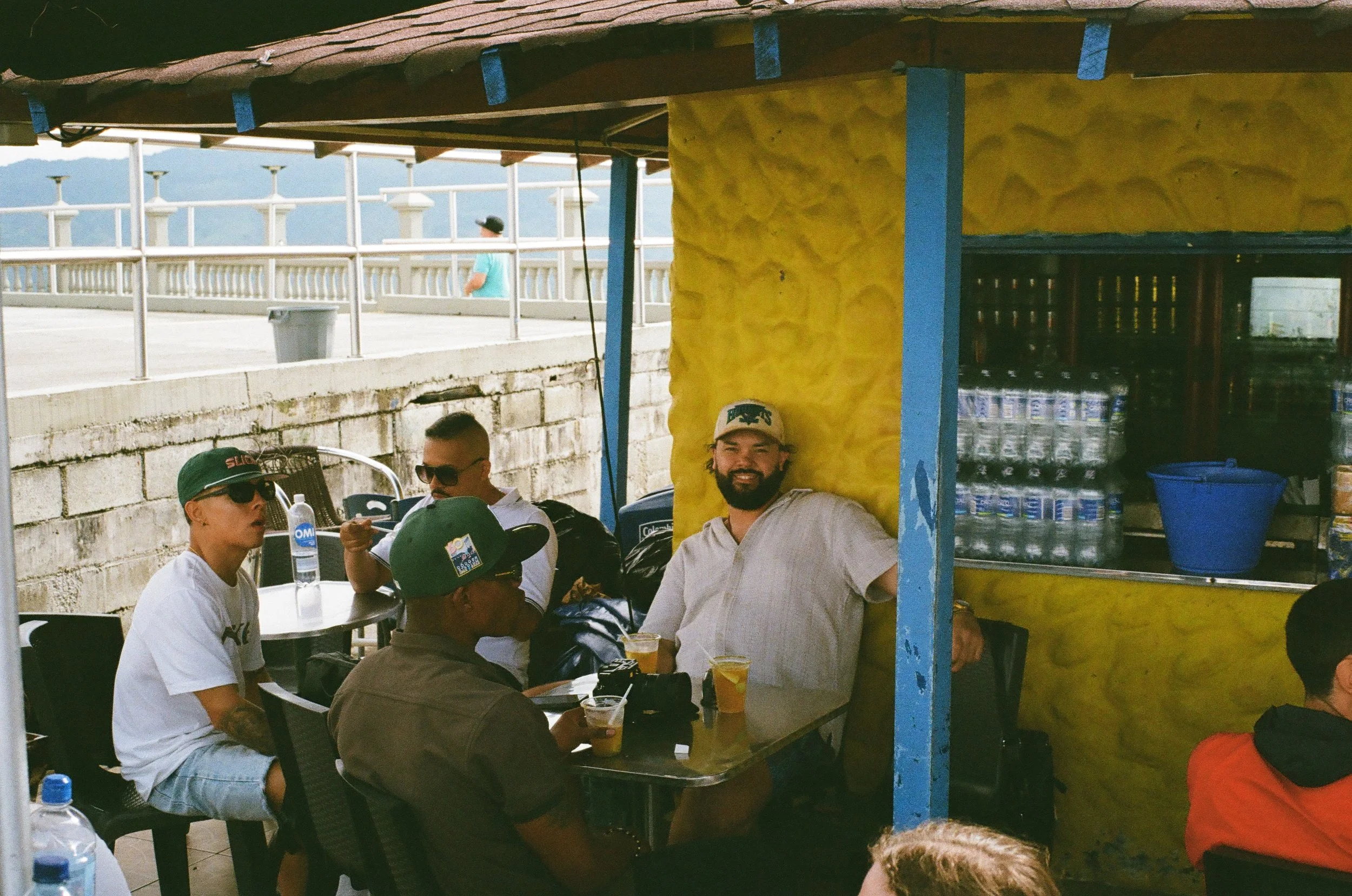 Group of men sitting at a booth under a yellow awning, enjoying drinks, with one man smiling at the camera in front of a bright yellow wall, near a snack stand with canned drinks and a blue bucket.