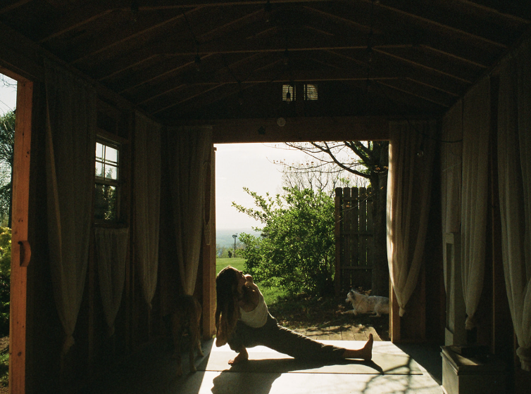 Person stretching inside a rustic shed with open doors, greenery outside, two dogs nearby.