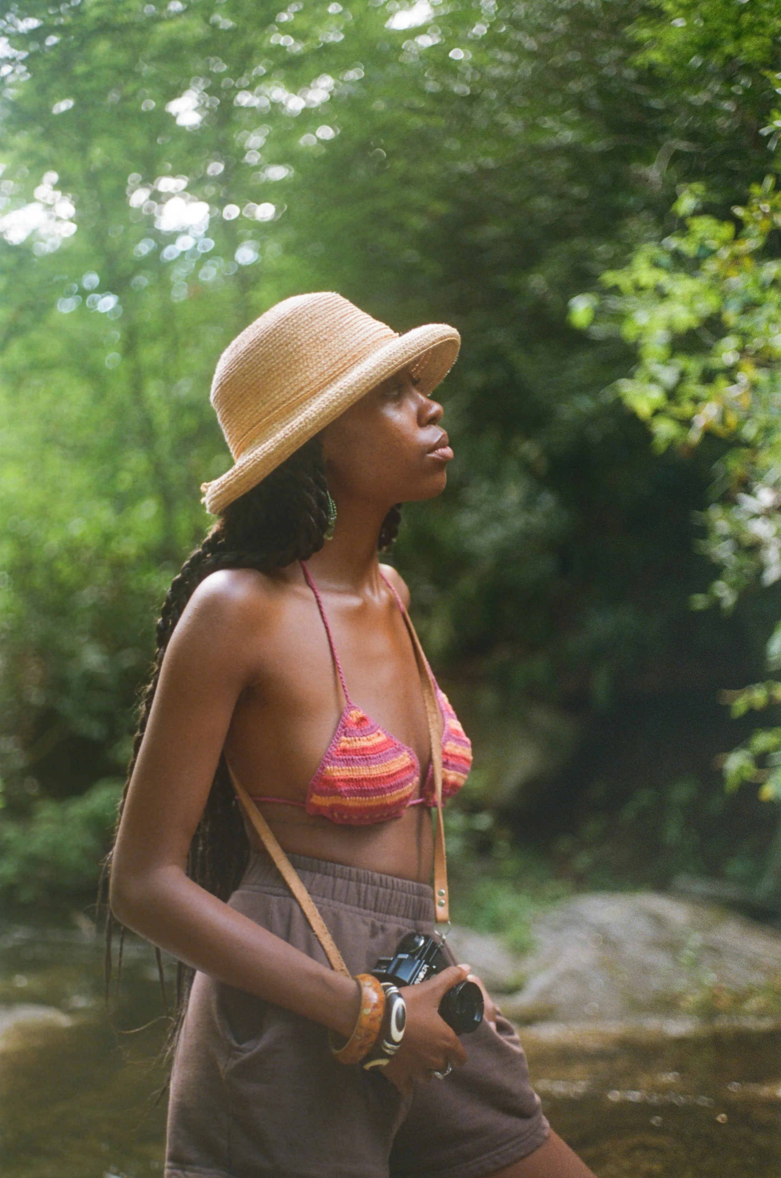 Woman wearing a straw hat and a colorful bikini top, holding a camera, standing outdoors in a green, wooded area.