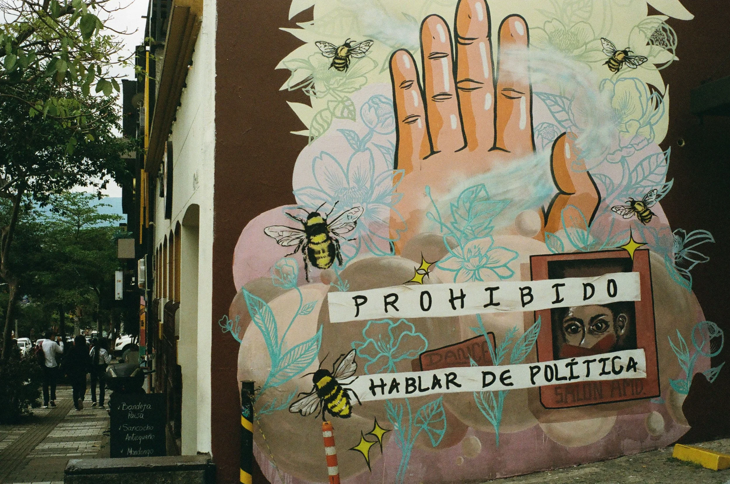 Street art mural depicting a hand with bees, flowers, and smoke, with a sign that reads 'Prohibido hablar de política' (Talking about politics is prohibited).