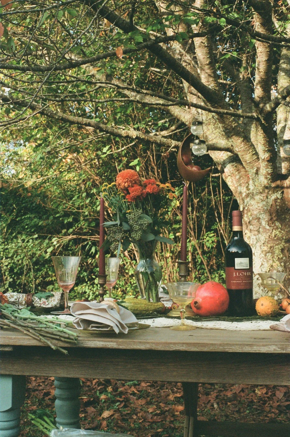 An outdoor table set with flowers, wine, candles, and decorative items under a large tree.