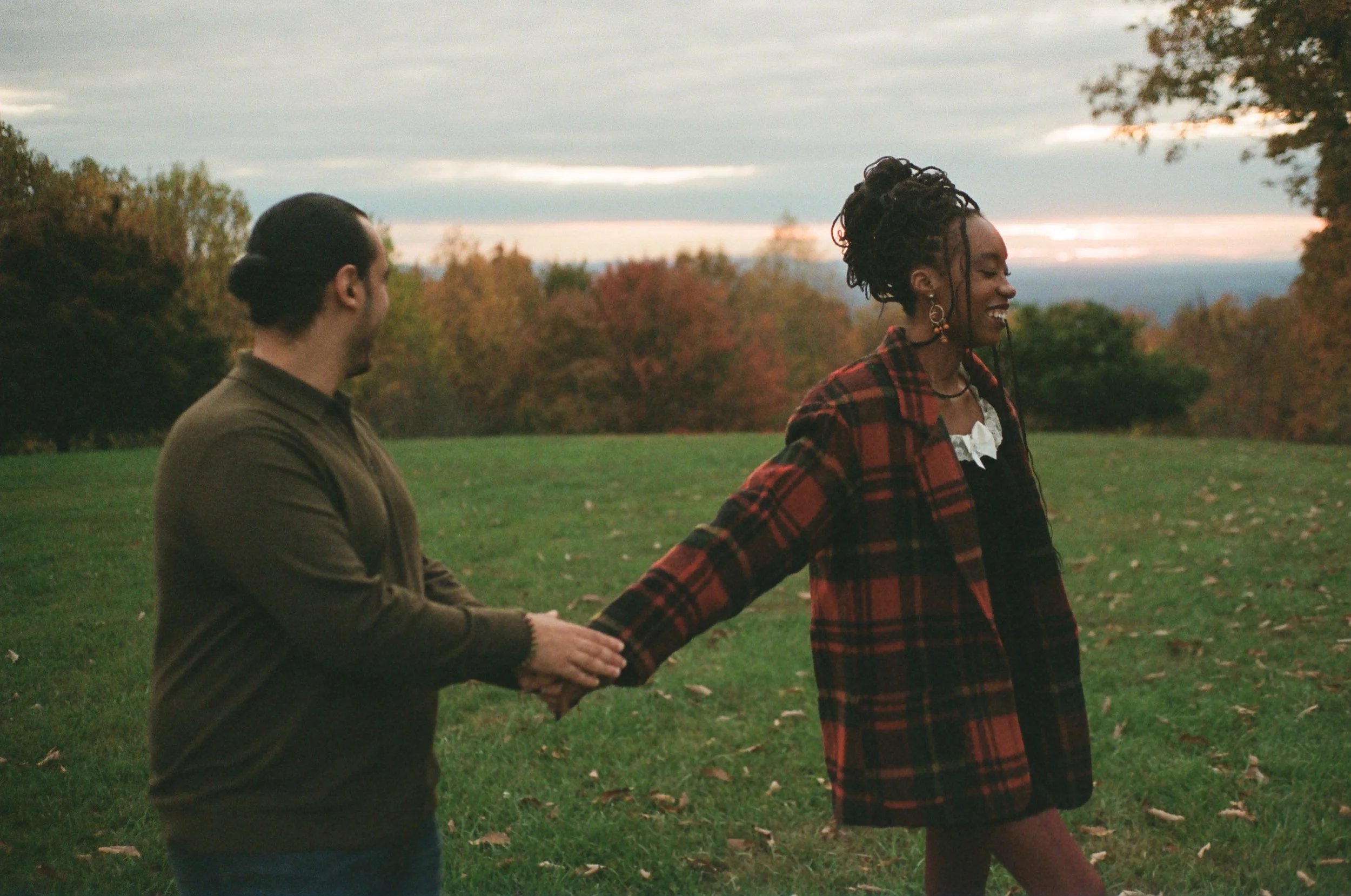 A man and woman holding hands and walking outdoors in a grassy park during sunset, with trees and clouds in the background.