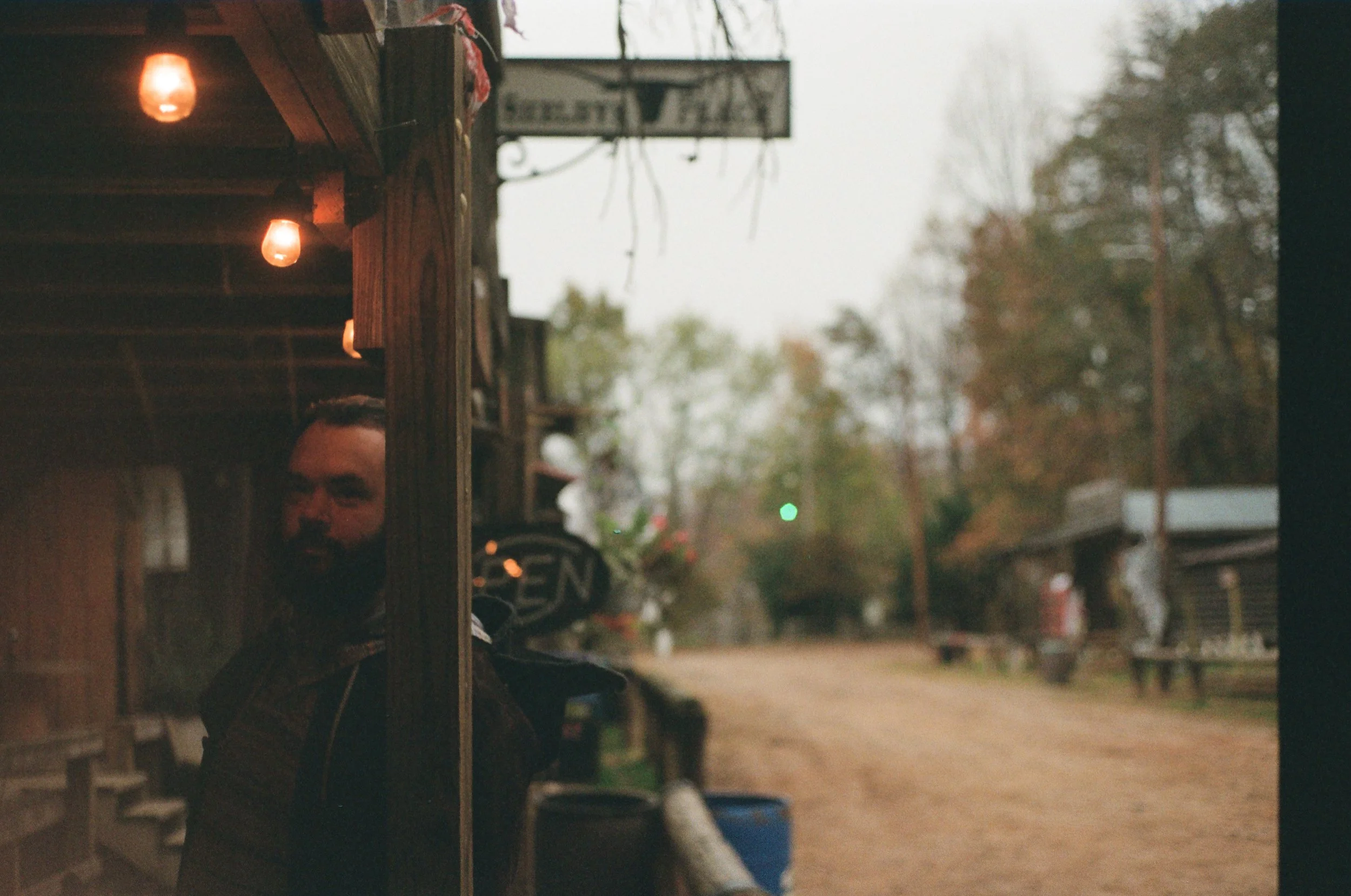 A man with a dark beard and hair tied back, standing outside a rustic building with warm string lights, on an overcast day with trees and a dirt road in the background.