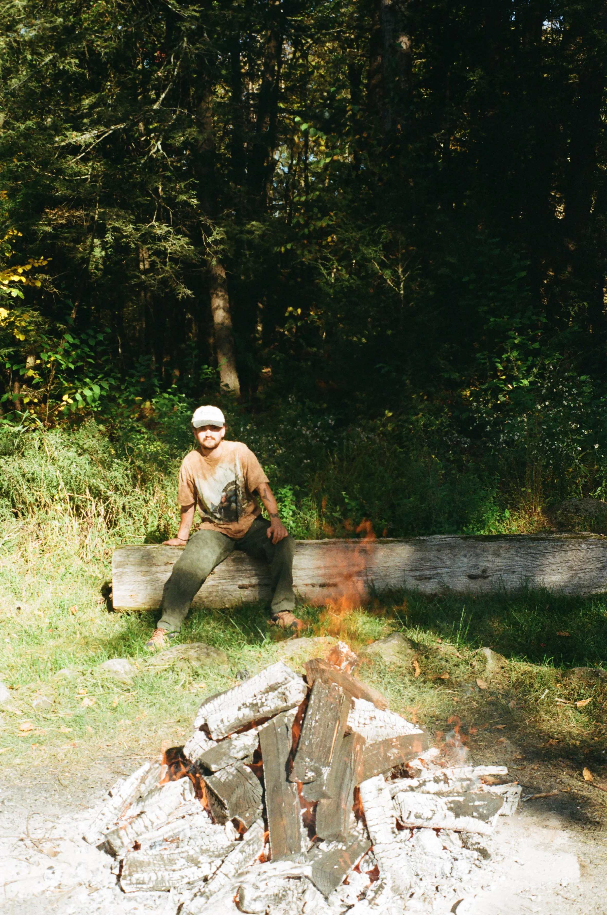 A man sits on a large log near a campfire in a forest with dense trees and green foliage.