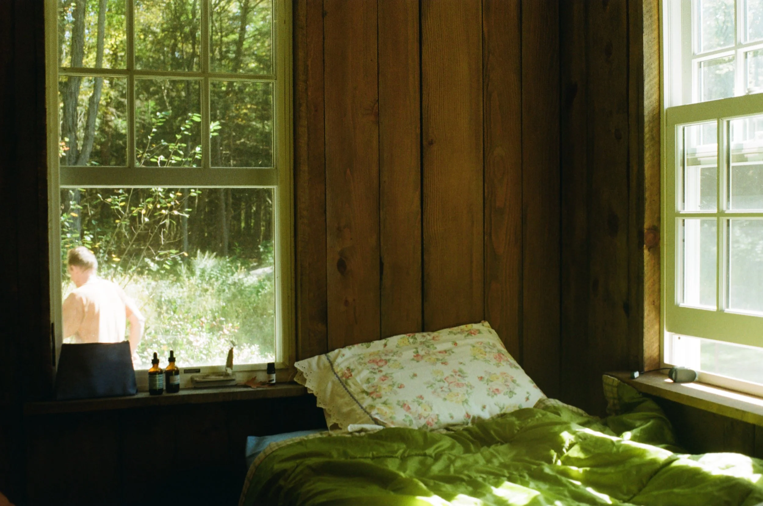 A rustic bedroom with wood-paneled walls, a bed with floral pillow and green bedding, and large windows showing outdoor greenery. A person is outside near the window.