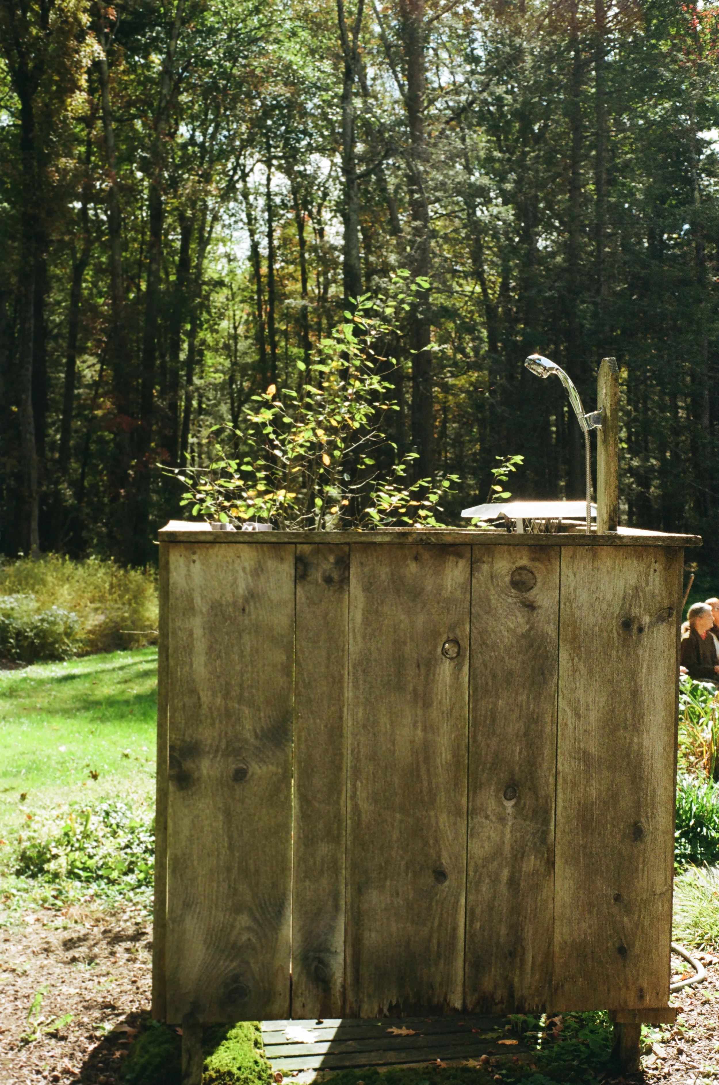 Wooden outdoor structure with a garden hose, faucet, and a sun umbrella, set in a backyard with trees and greenery.