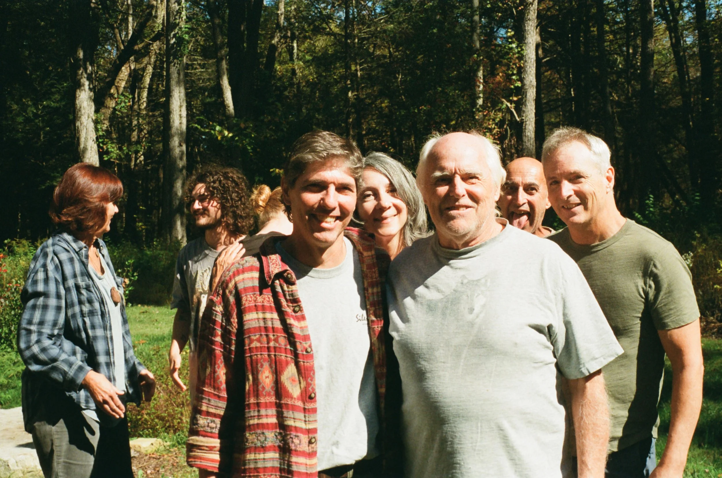 Group of six adults outdoors in a wooded area, smiling and posing for the camera.
