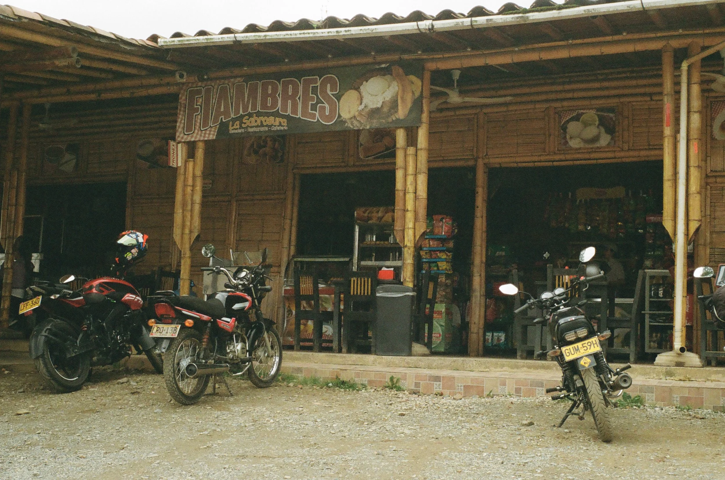 A small rustic restaurant with a bamboo exterior and a sign that says 'FIAMBRES La Sabrosura'. In front, there are two motorcycles parked on a dirt ground.