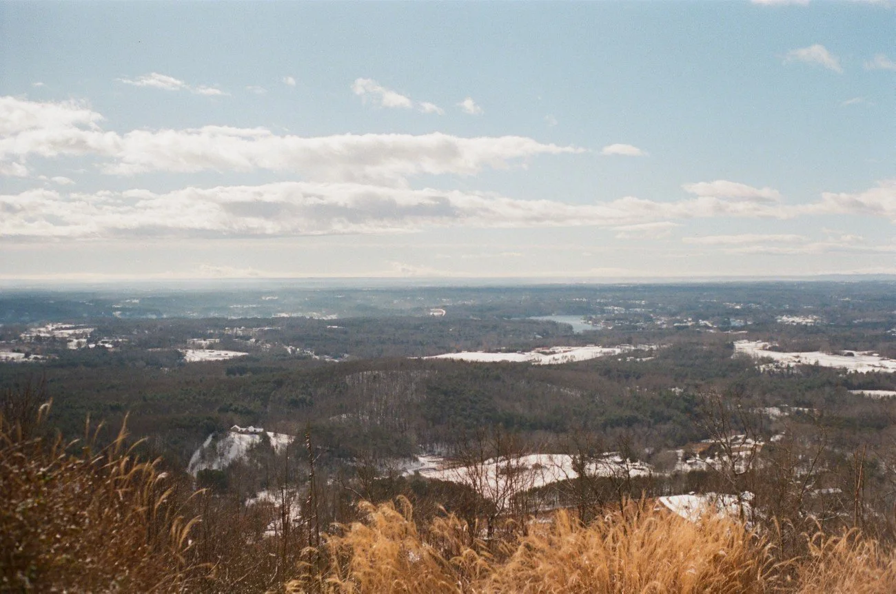 View of a landscape with trees, snow patches, and a distant lake or river under a partly cloudy sky.