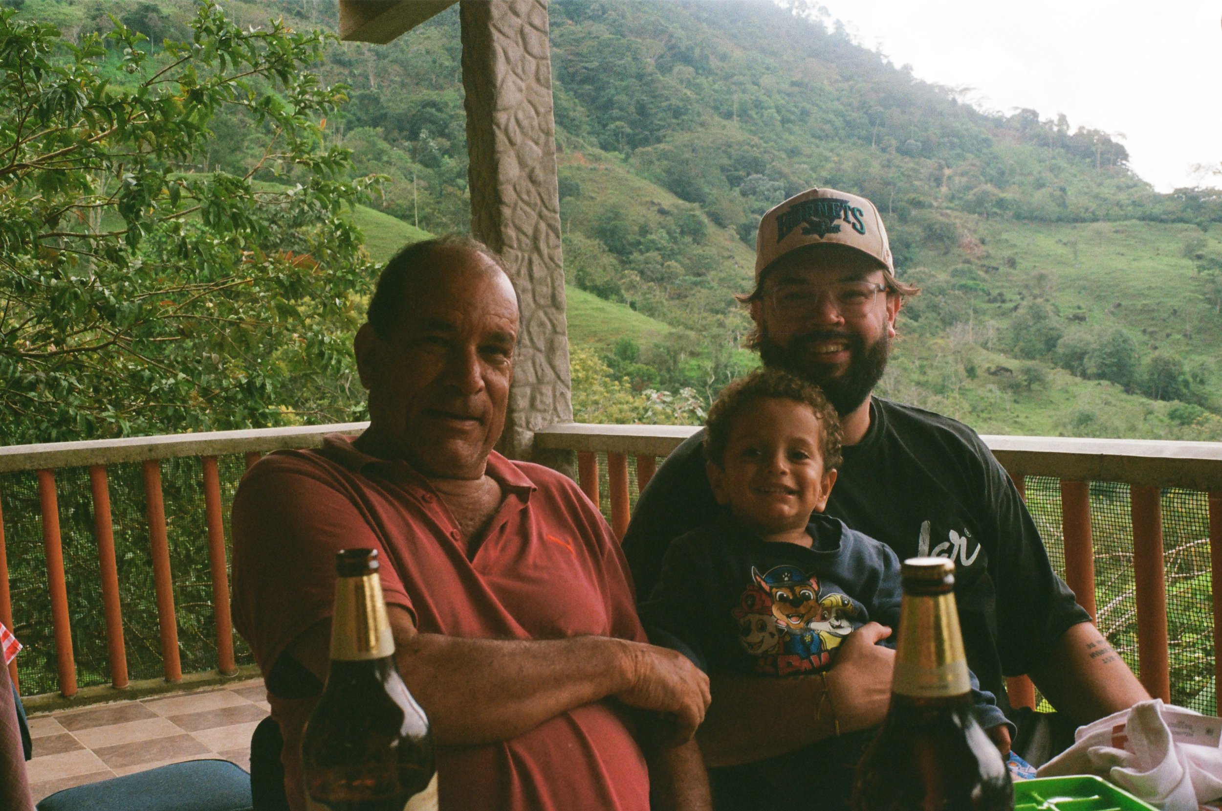 Three people, an elderly man, a middle-aged man, and a young boy, sit at a table on a porch overlooking a lush, green hillside with trees and vegetation. They are smiling, and there are bottles and a small green container on the table.