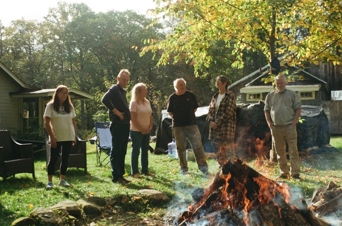 Six people standing outdoors around a campfire in a backyard, with trees and a house in the background during daytime.