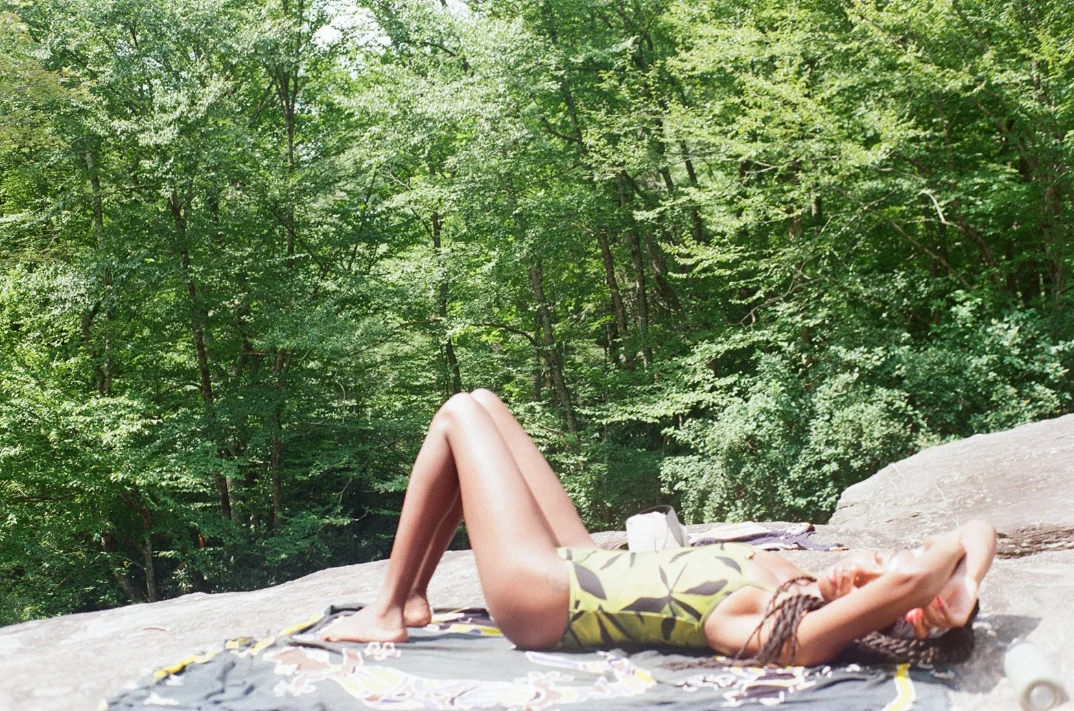 A woman lying on her back on a rock surface, sunbathing with her hands behind her head, in a forested area with lush green trees.