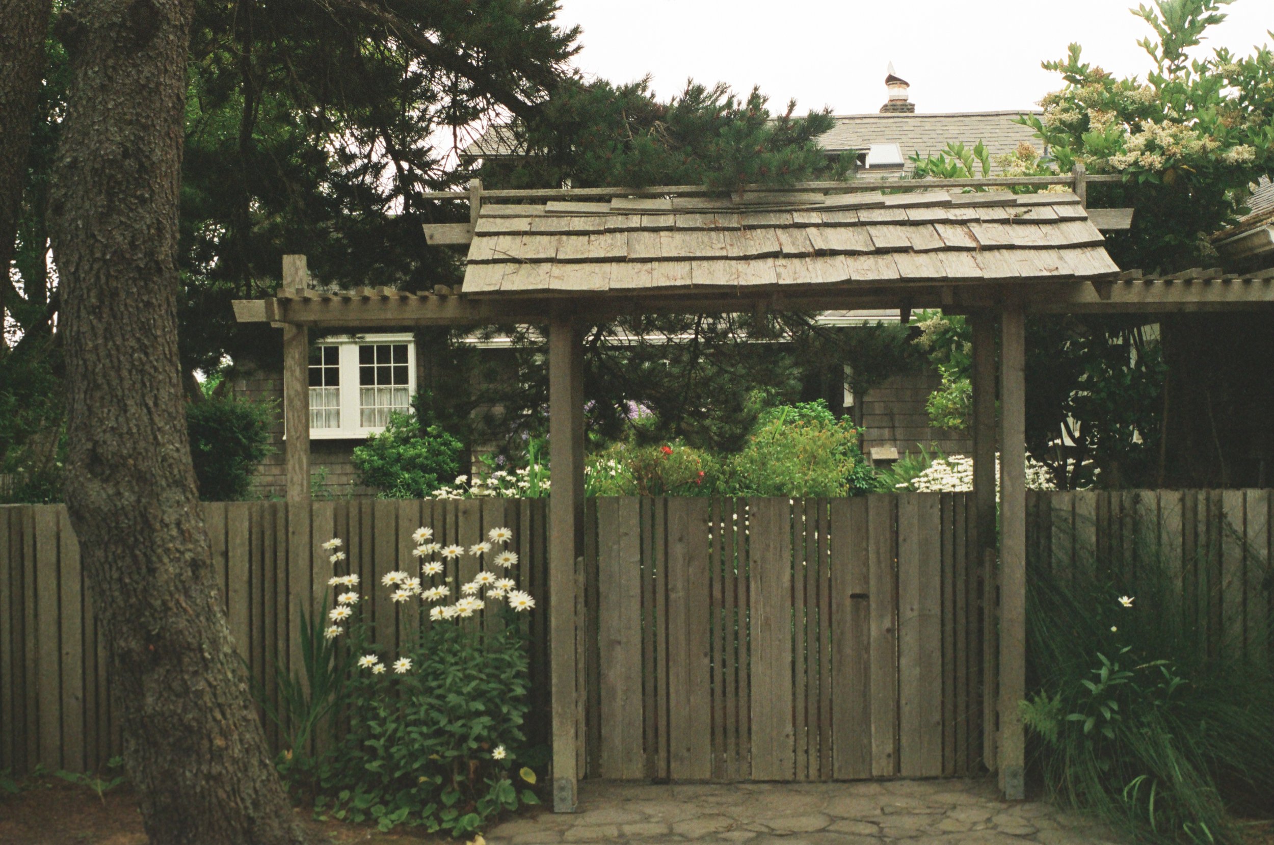 A wooden garden gate with a gatehouse structure on top, surrounded by trees, flowering plants, and a rustic house in the background.