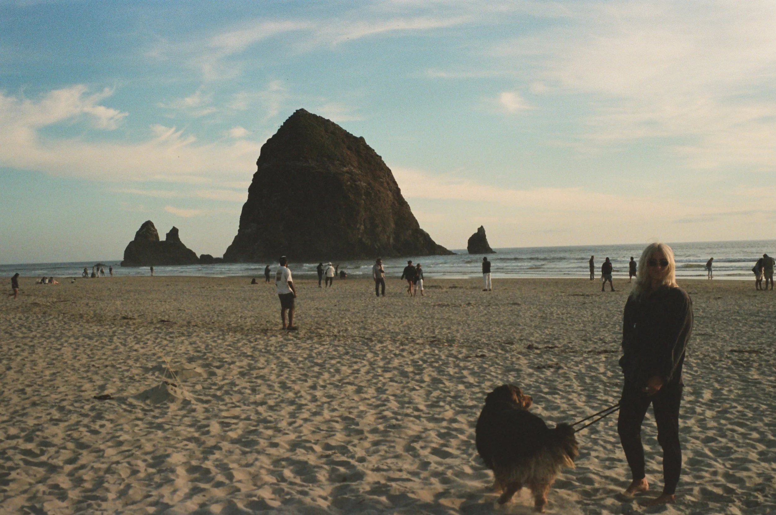 A woman with sunglasses walking a dog on a sandy beach during sunset, with large rocks and a sea stack in the background.