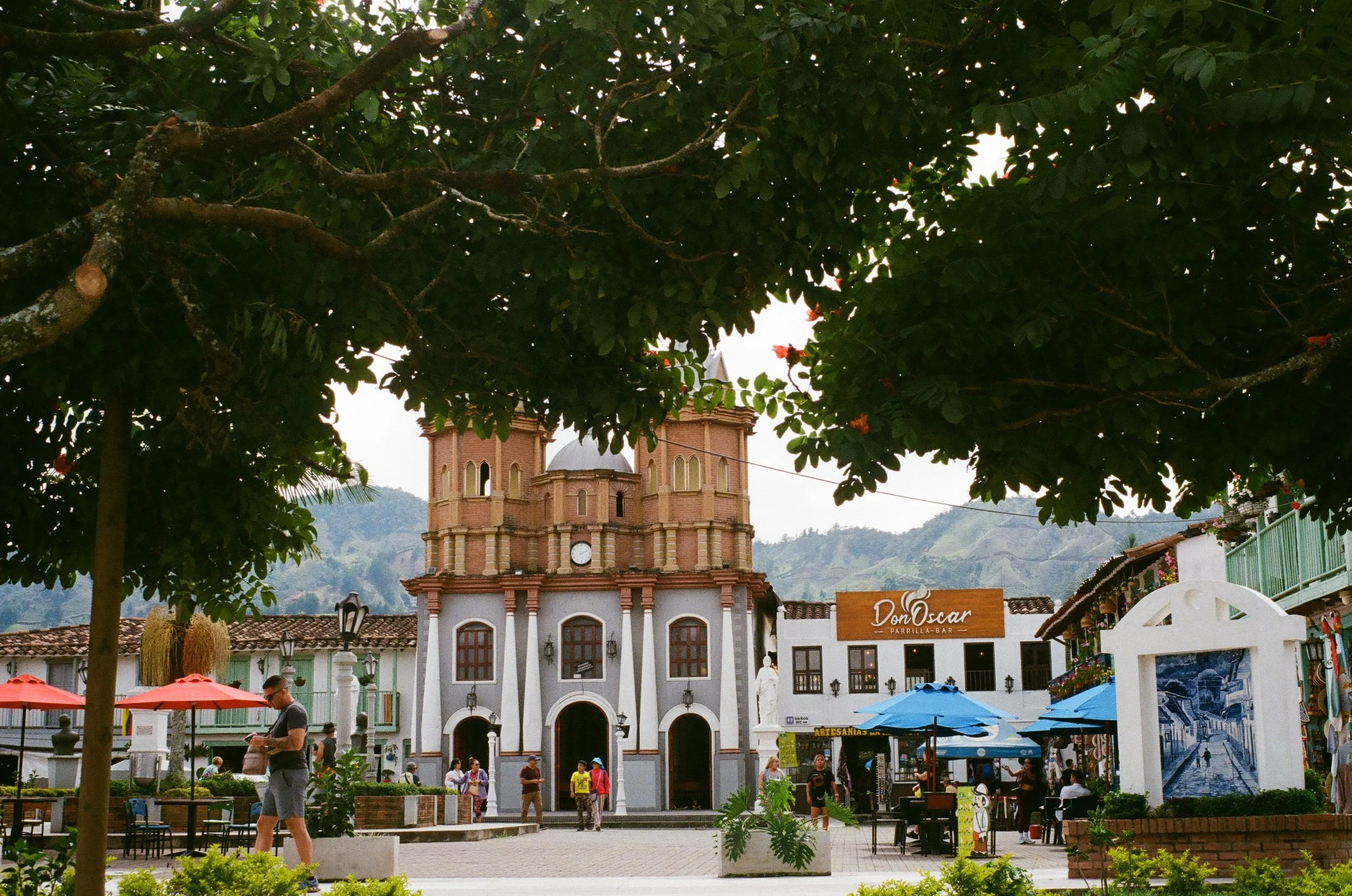 A scenic town square featuring a large brick church with twin towers and a domed roof, surrounded by shops and cafes with colorful umbrellas, in a mountainous landscape.