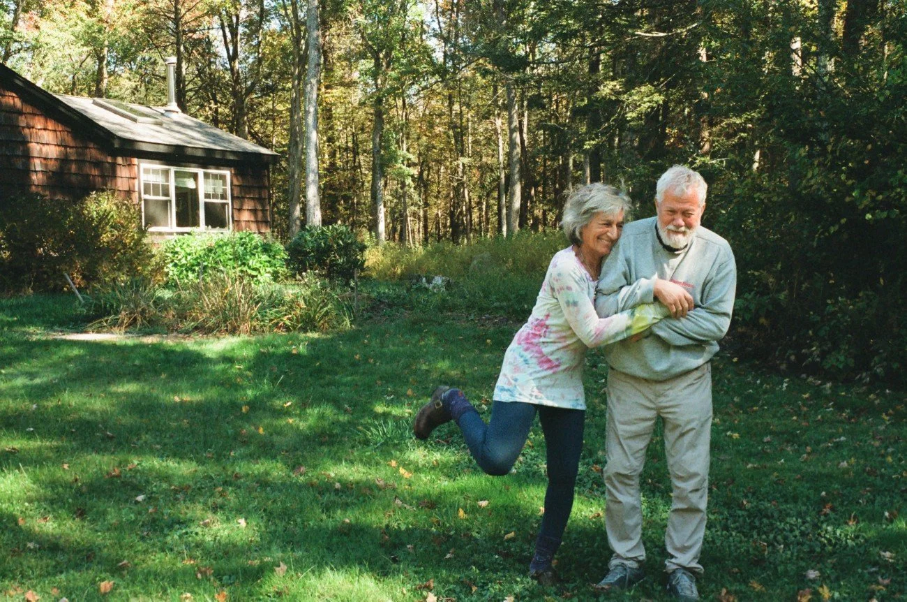 An elderly woman and man are outdoors happily playing together on a grassy yard, with a small wooden house and trees in the background.