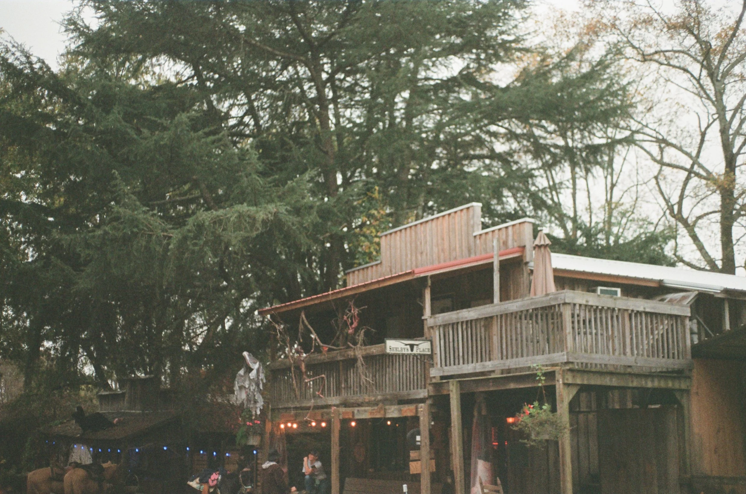 A wooden house with a second-story balcony, surrounded by trees, with string lights and people beneath it.