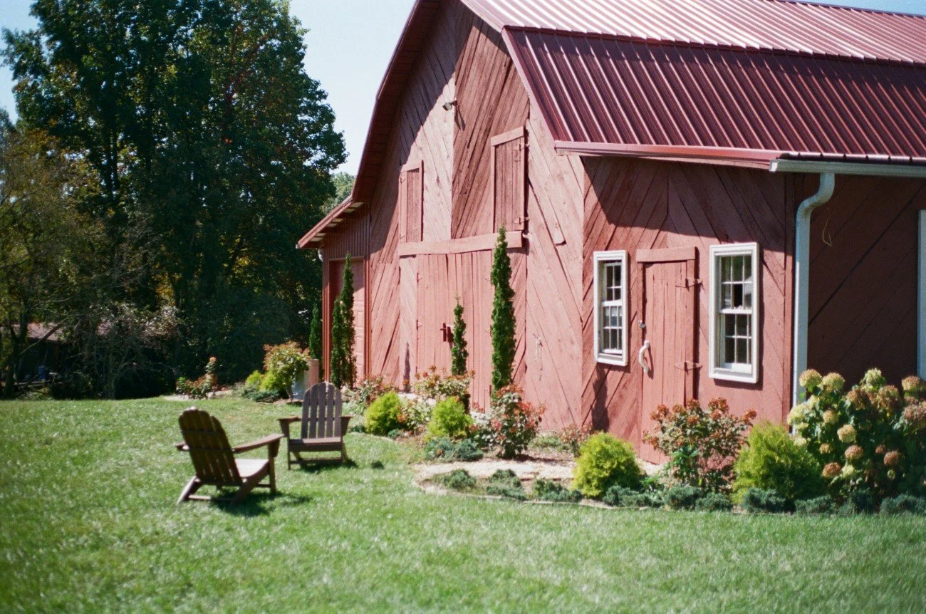 Red barn with white-framed windows, surrounded by a landscaped garden with green grass, small shrubs, and tall trees in the background.