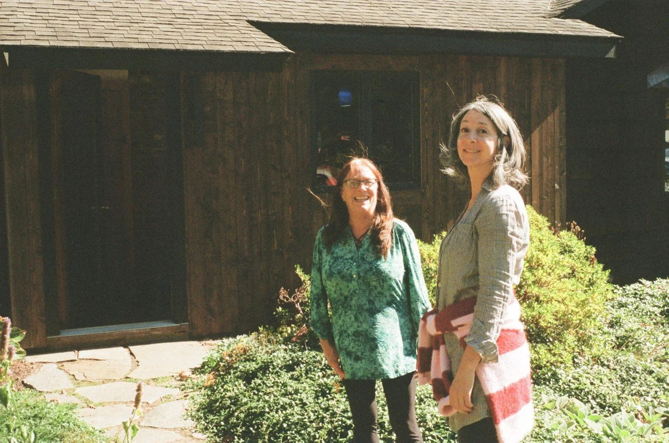 Two women standing outside in front of a dark wooden house, smiling, with greenery and garden plants around them.