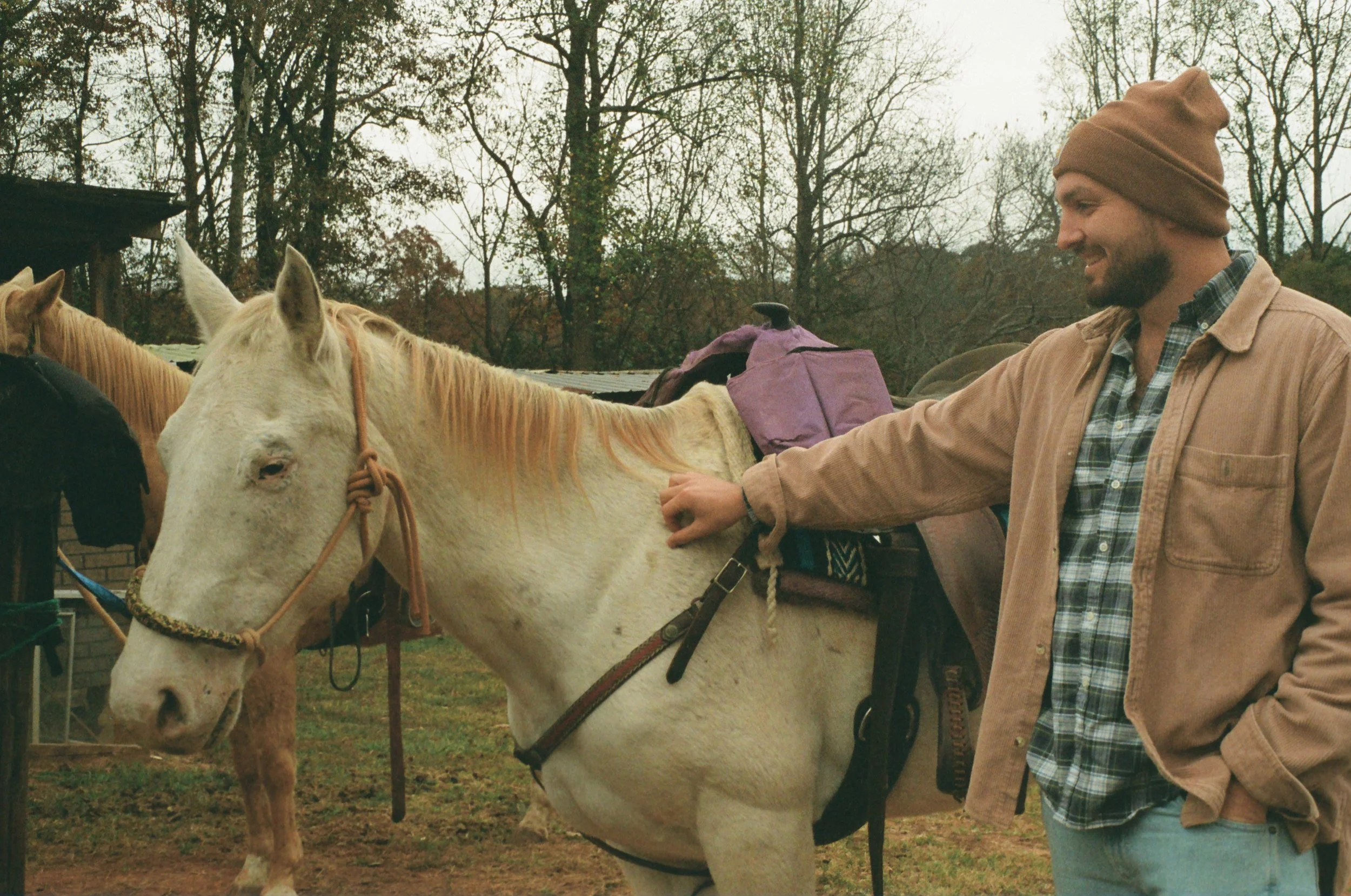 A man in a brown beanie and beige jacket petting a white horse with a saddle and blanket, outdoors on a cloudy day with trees in the background.