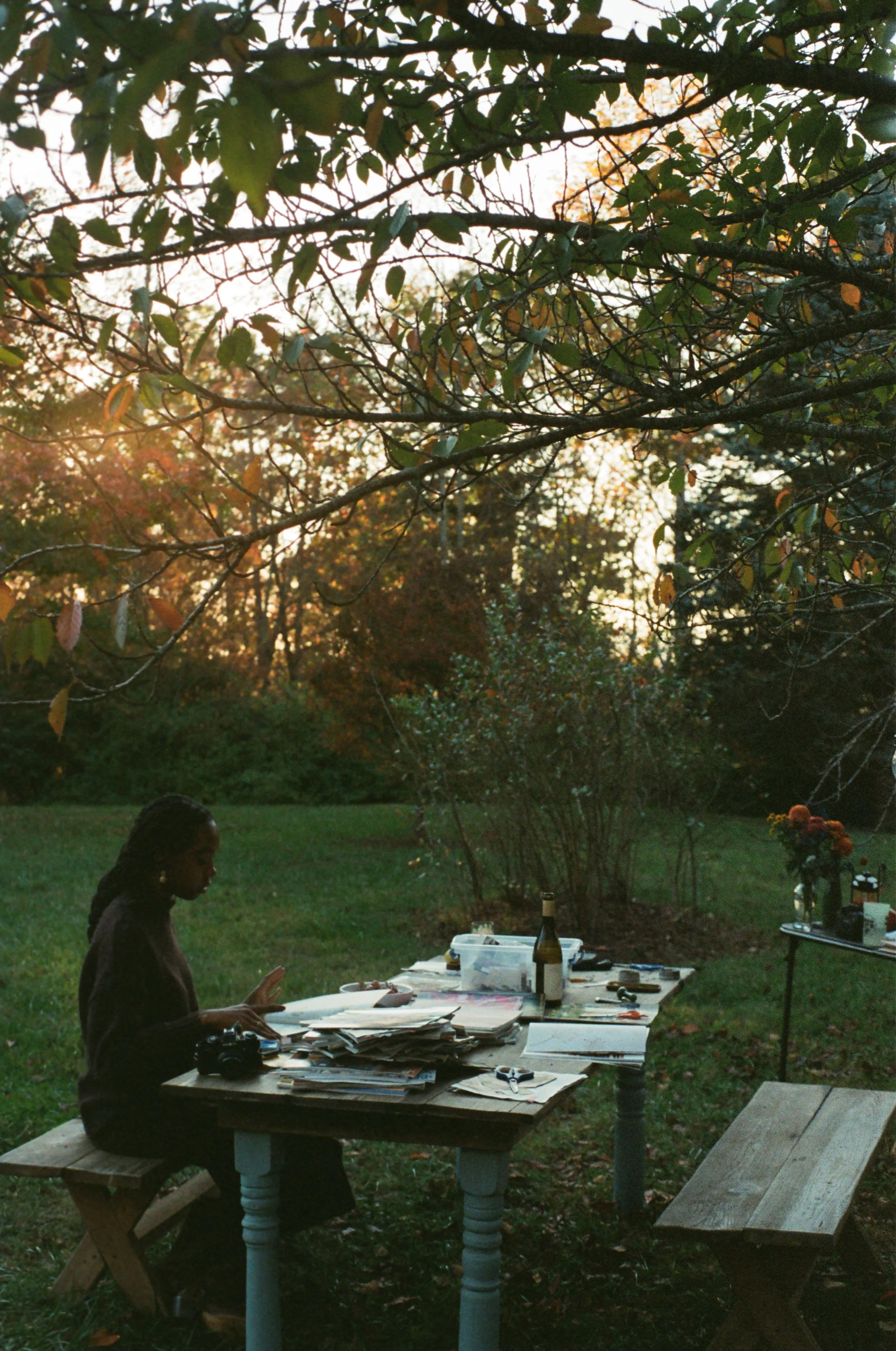 Woman sitting at a table outdoors in a garden during sunset, surrounded by books or papers, with a camera and a bottle on the table, and another table with flowers in the background.