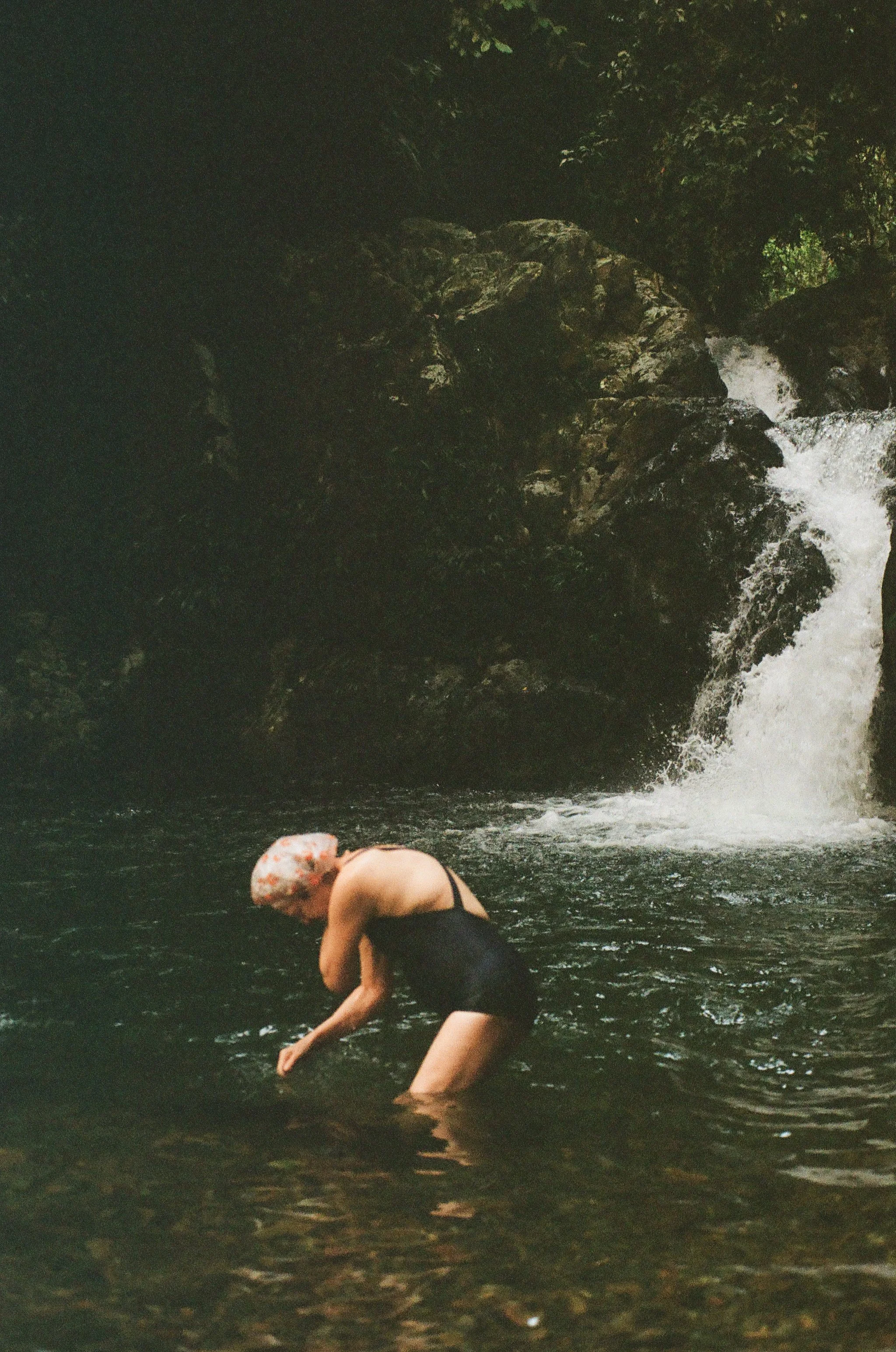 A person with a bald head, wearing a black swimsuit, standing in a body of water near a waterfall and rocky landscape.