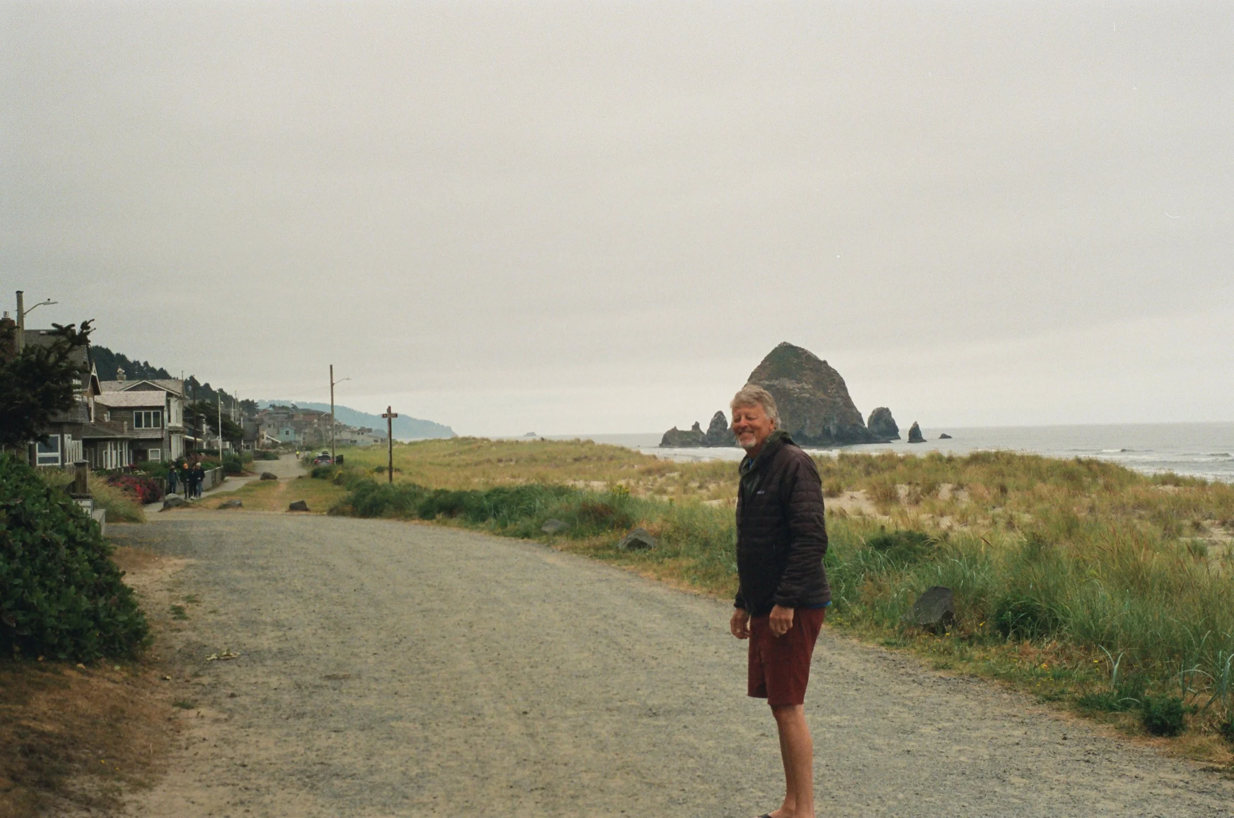 A man standing barefoot on a gravel road near the beach, with houses on the left and a large rock formation in the ocean in the background on an overcast day.
