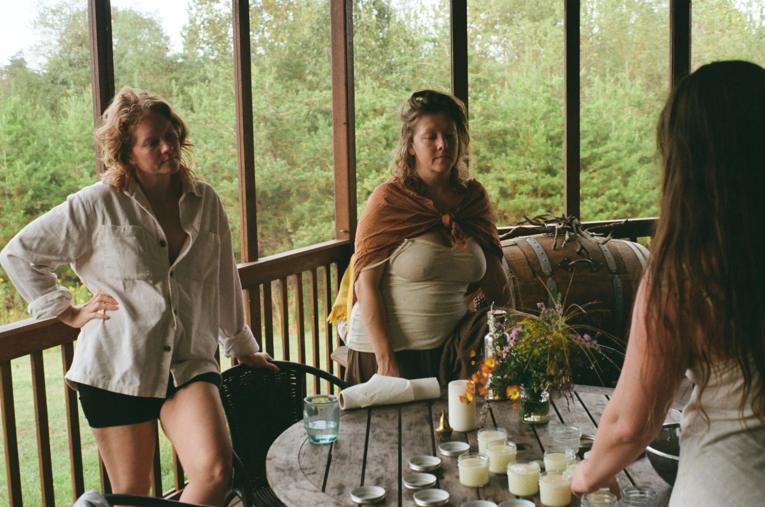 Three women gathered around a wooden table on a screened porch, with candles and flowers, during daytime. The background shows trees and greenery.