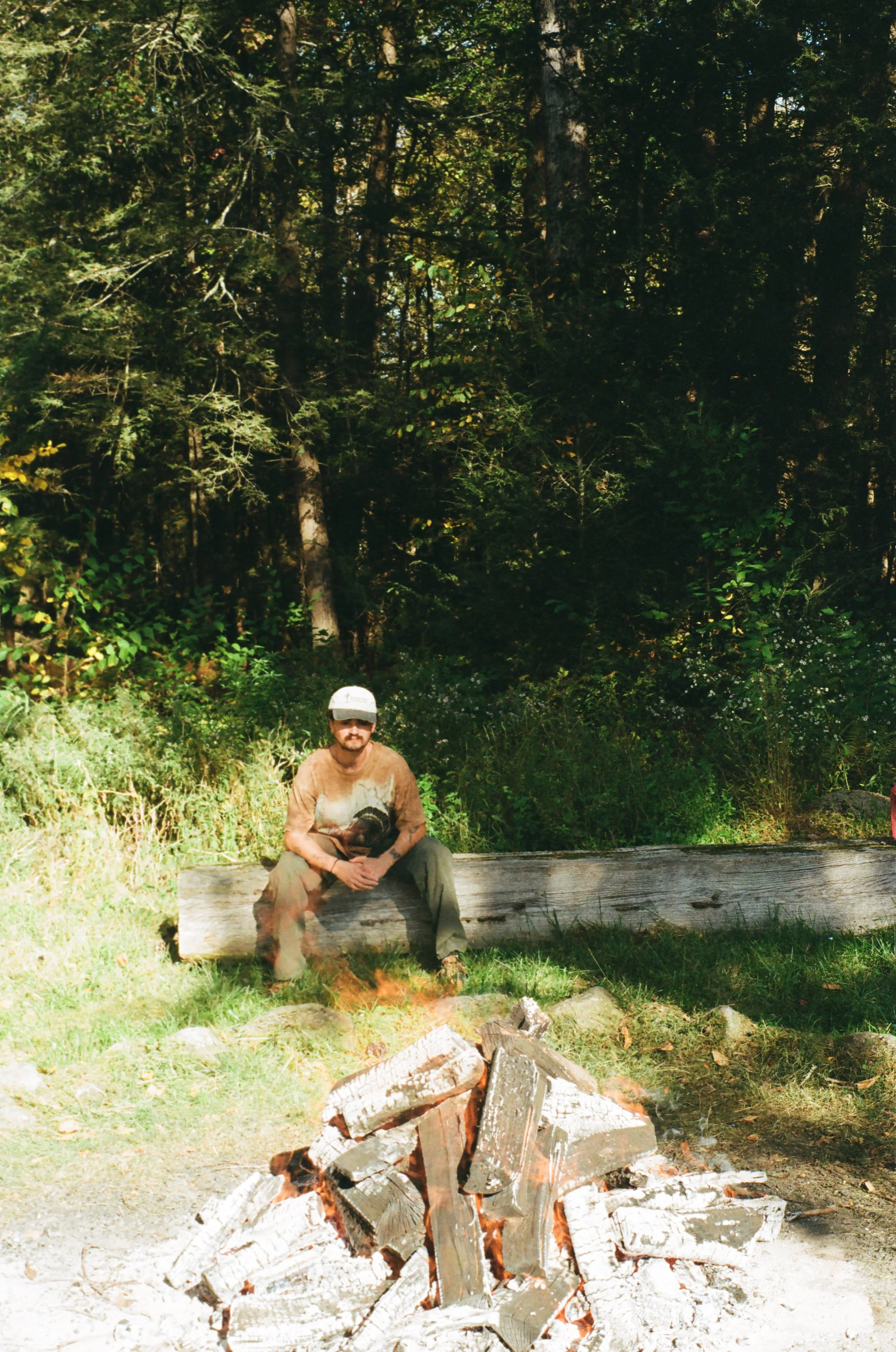 A man sitting on a large log in a forest clearing near a campfire with wood logs burning on the ground, surrounded by tall trees and green foliage.
