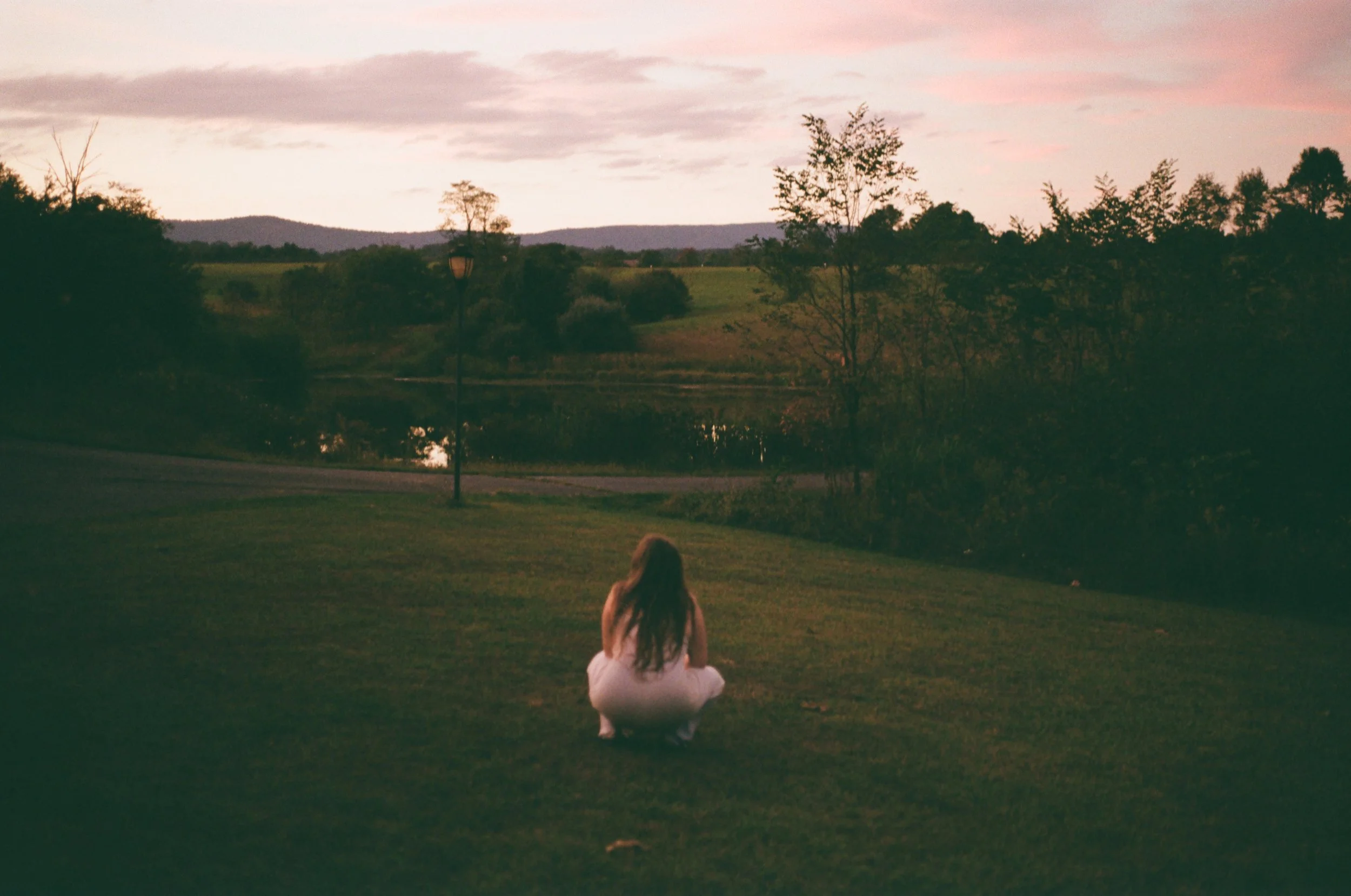 A woman with long hair squatting on grass near a lake during sunset, with trees and distant hills in the background.