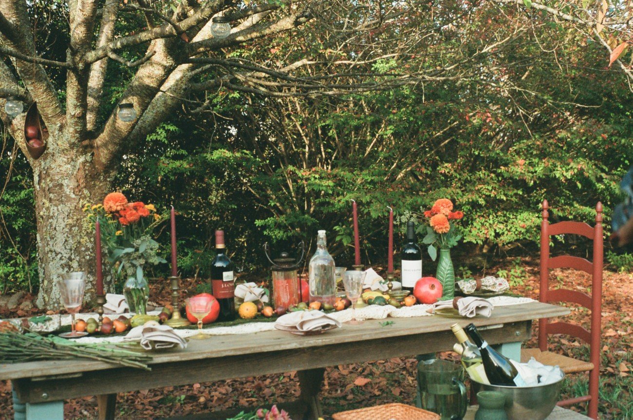 Outdoor dinner table decorated with flowers, wine bottles, fruits, candles, and glasses, set in a garden with trees and fallen leaves.