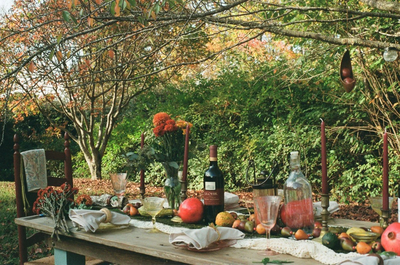 An outdoor table set for a meal in a garden with trees and green foliage. The table has a lace table runner, candles, wine, glasses, flowers, and various fruits.