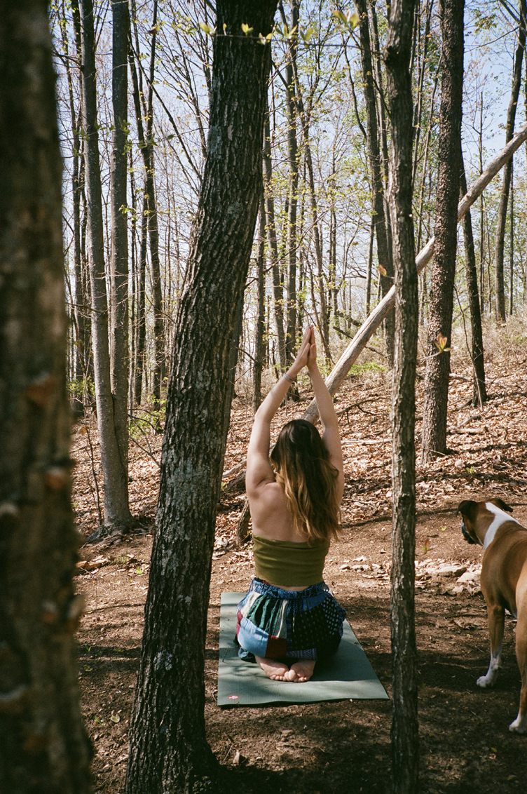 A woman practicing yoga outdoors in a forest, kneeling on a yoga mat with arms raised above her head in a prayer position, accompanied by a dog standing nearby.