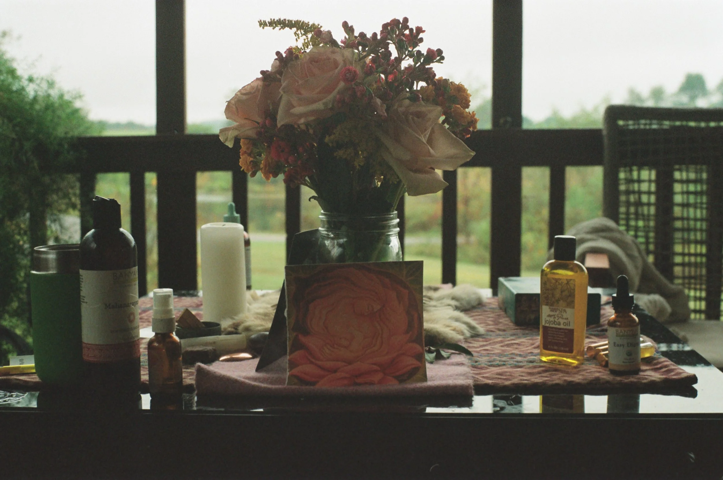 A table with a floral centerpiece, various bottles of essential oils, a candle, a card, and a piece of fabric or cloth, set outdoors on a porch with a view of greenery outside.