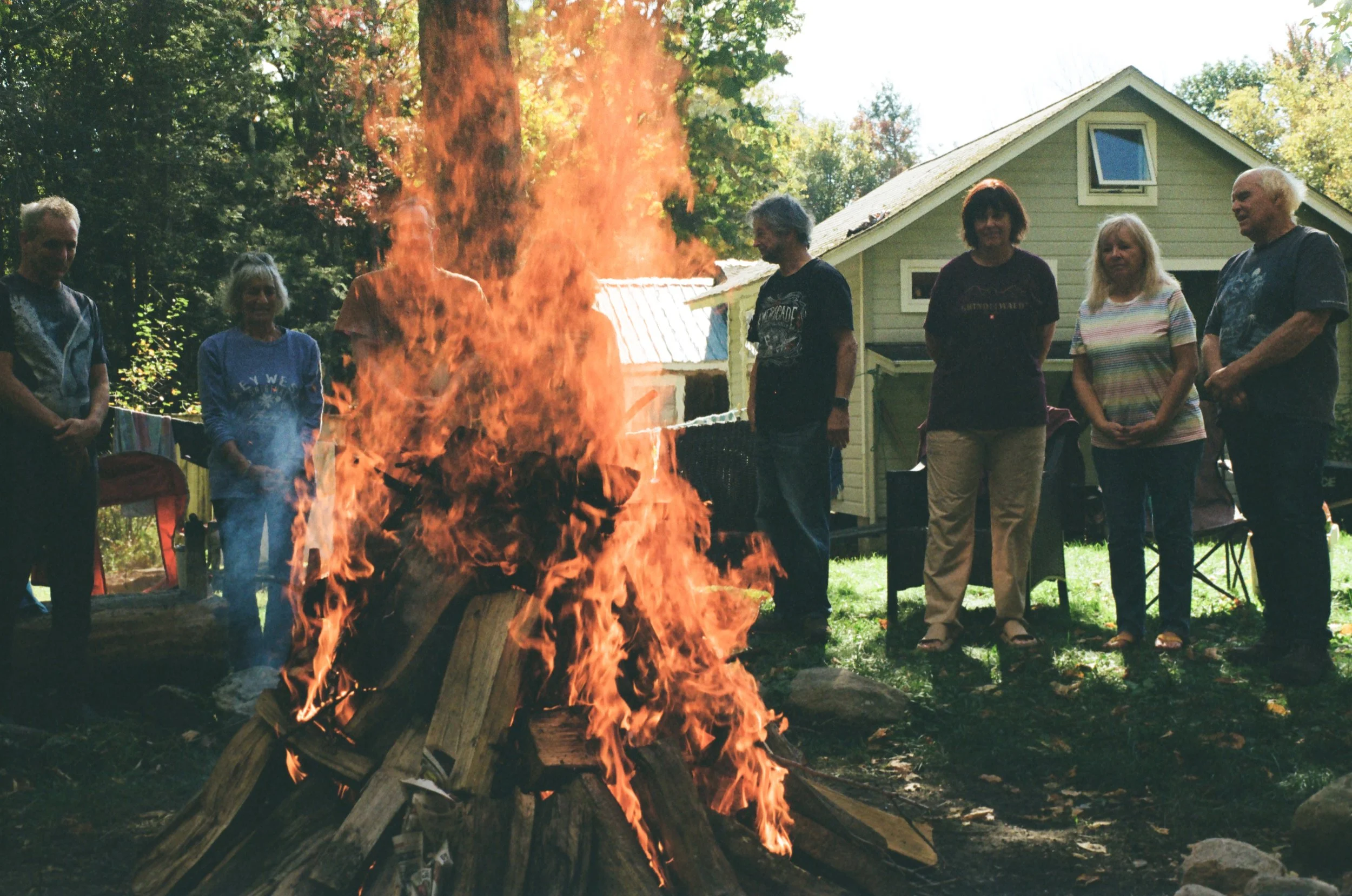 A group of seven people standing around a large outdoor fire with burning logs, in a backyard setting with trees and a house in the background.