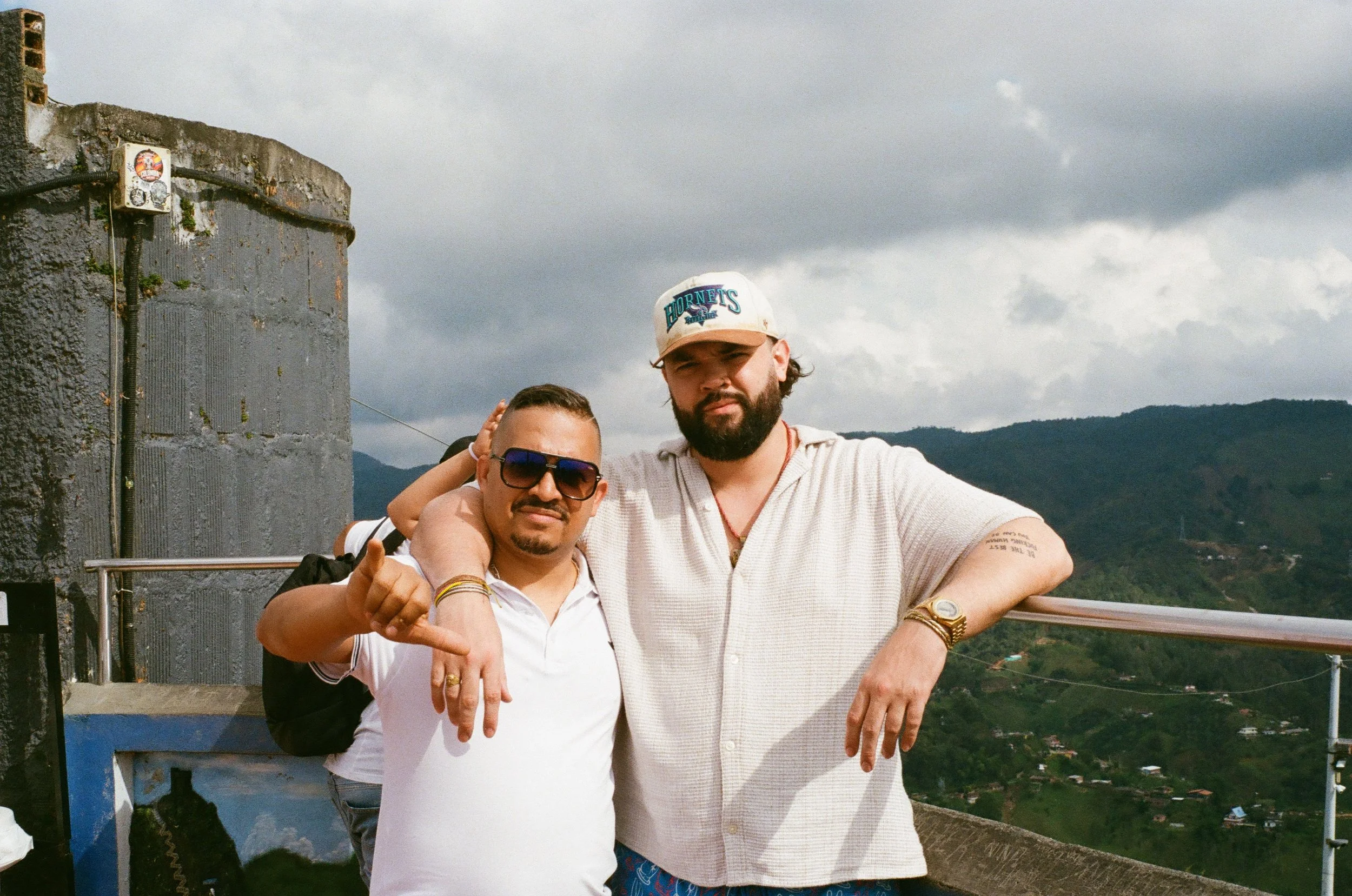 Two men stand together on a mountain viewing platform with a rural landscape in the background. The man on the left wears sunglasses and a white polo shirt, while the man on the right wears a white cap, a light-colored, short-sleeved shirt, and has a