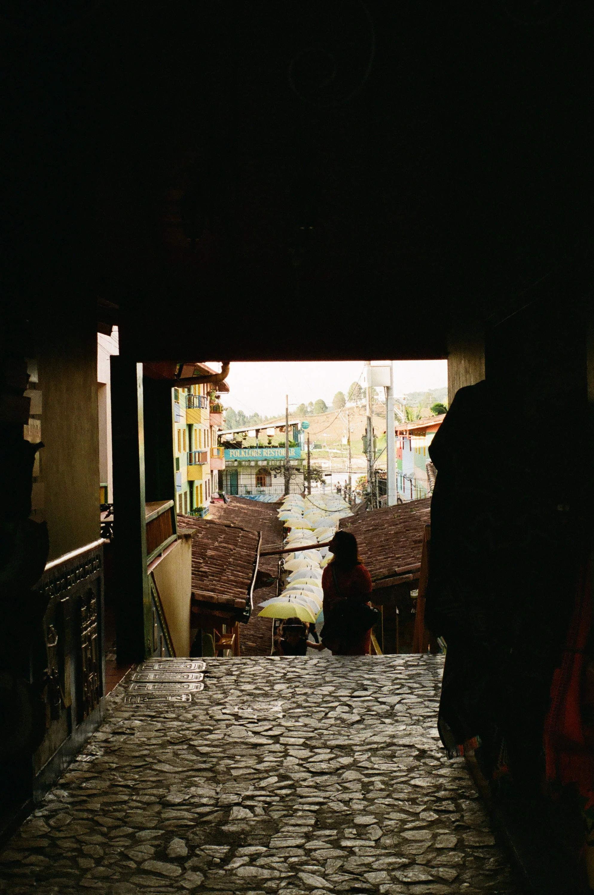 View from inside a dark building looking out onto a colorful street with umbrellas and people, including a woman with a backpack.