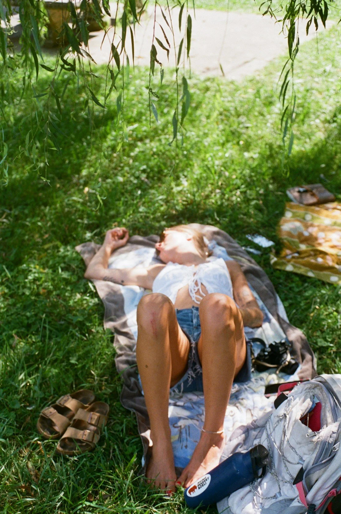 Person lying on a blanket under a tree on a grassy area, with belongings around, appearing to sleep or rest.