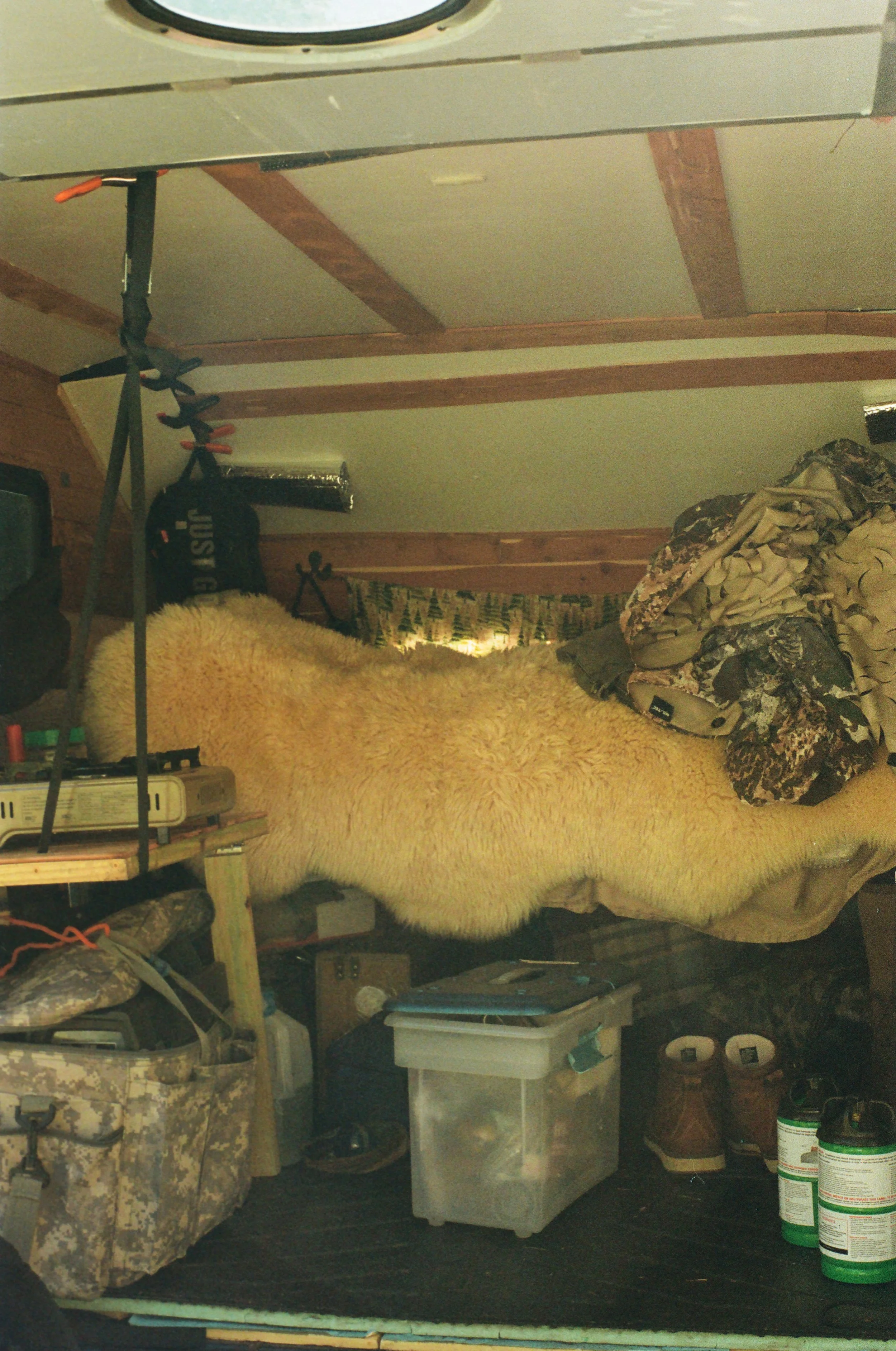 Inside a small cabin or camper with a bed, covered by a large white fluffy throw, and various camping gear and supplies around. There are jars, boots, plastic containers, and camouflage clothing.