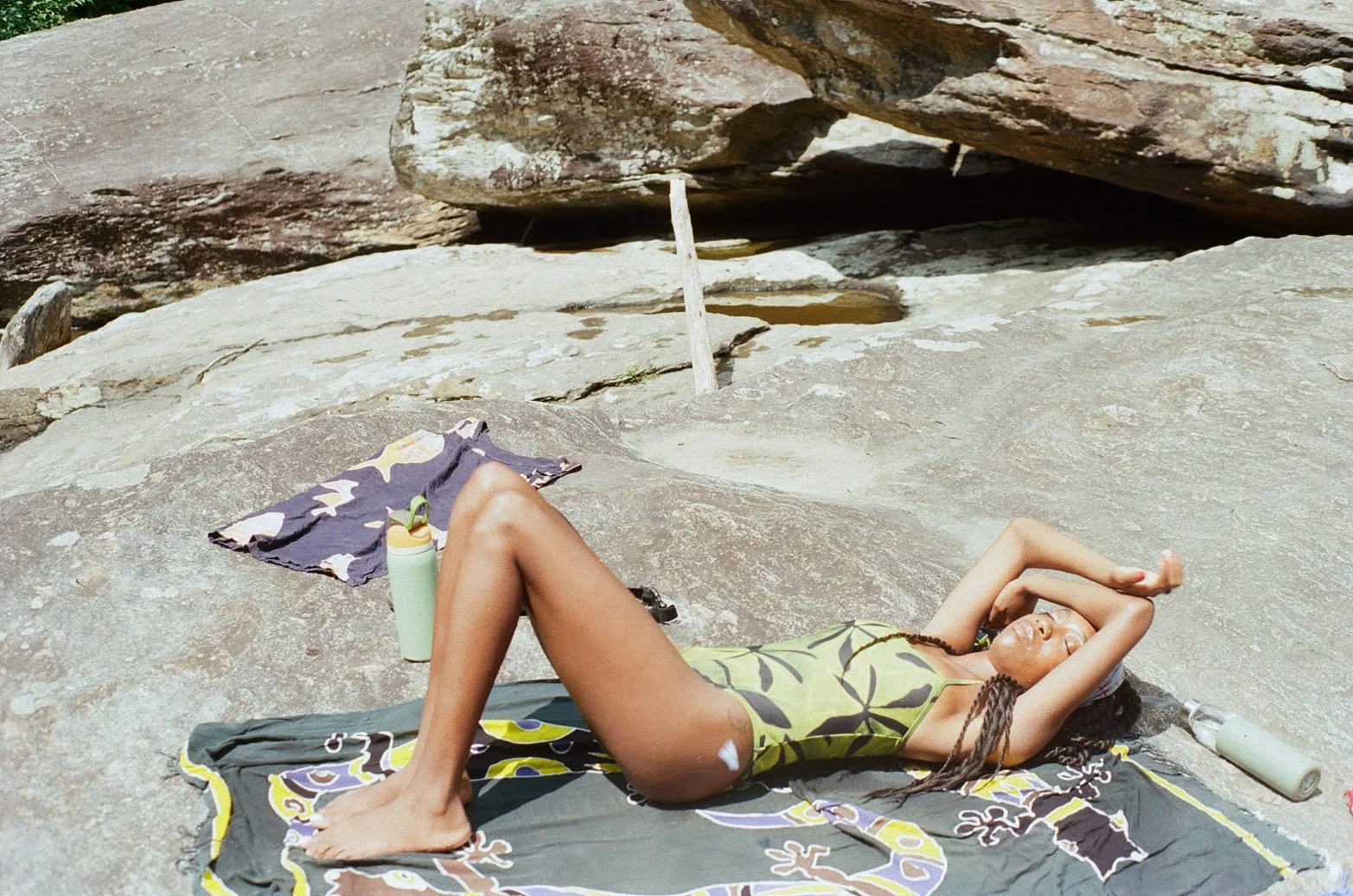A woman with long braids and sunglasses is lying on her back on a towel on a rocky surface, with her arms behind her head, near a large rock formation.