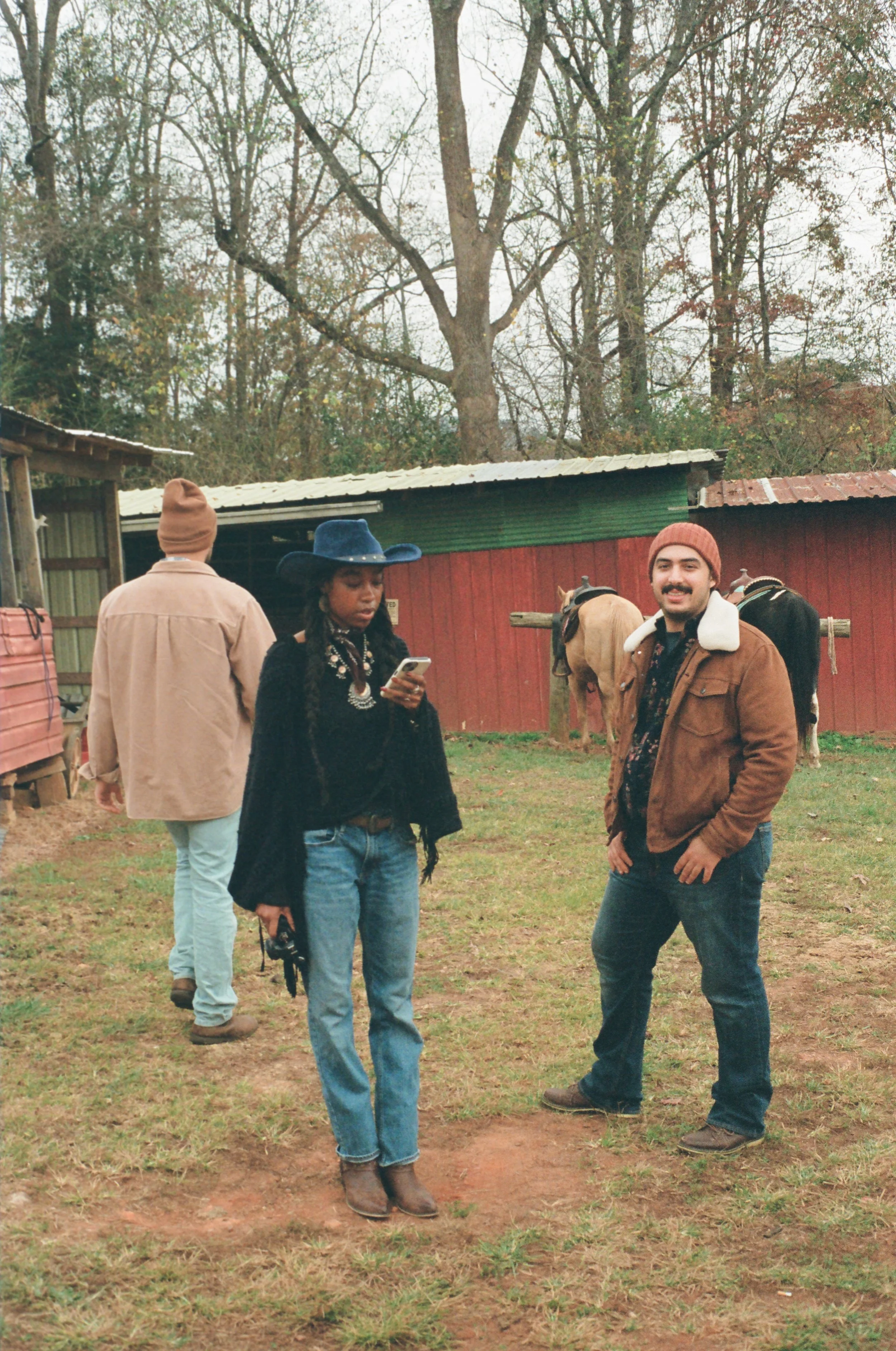 Three people on a farm, two standing and one walking away, with horses tied to a wooden fence and trees in the background.