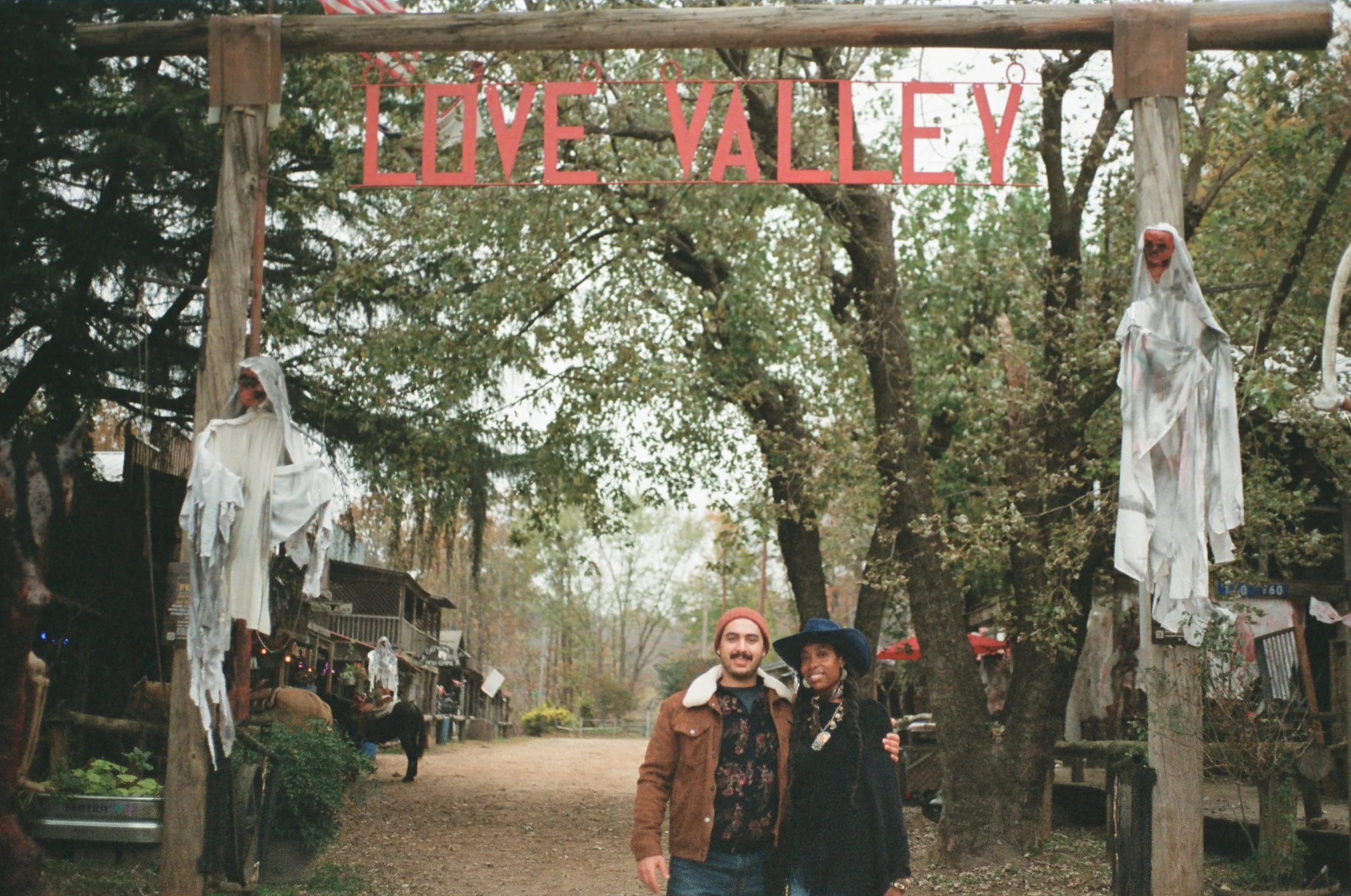 A man and woman standing together under a sign that reads 'Love Valley', surrounded by trees and decorations, with two ghostly figures hanging from posts on either side.