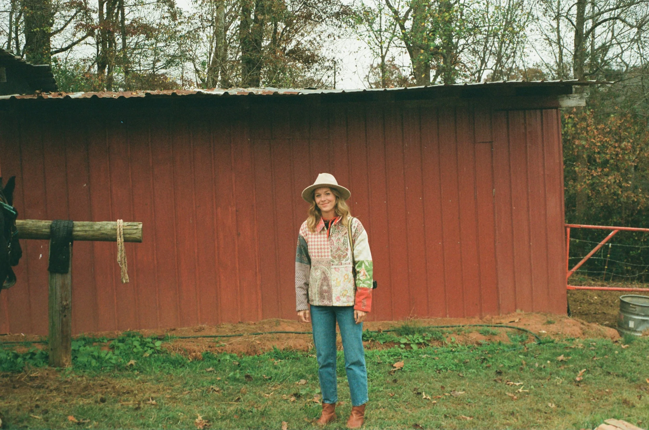 A woman standing outdoors in front of a red barn wearing a wide-brimmed hat, a colorful patchwork jacket, blue jeans, and brown boots.