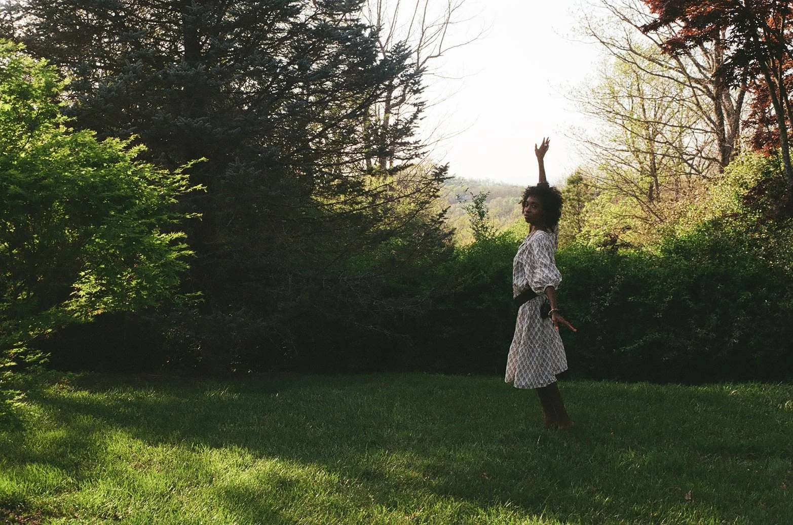 A woman standing on grass in a sunny outdoor setting, surrounded by trees and bushes, with one arm raised above her head.