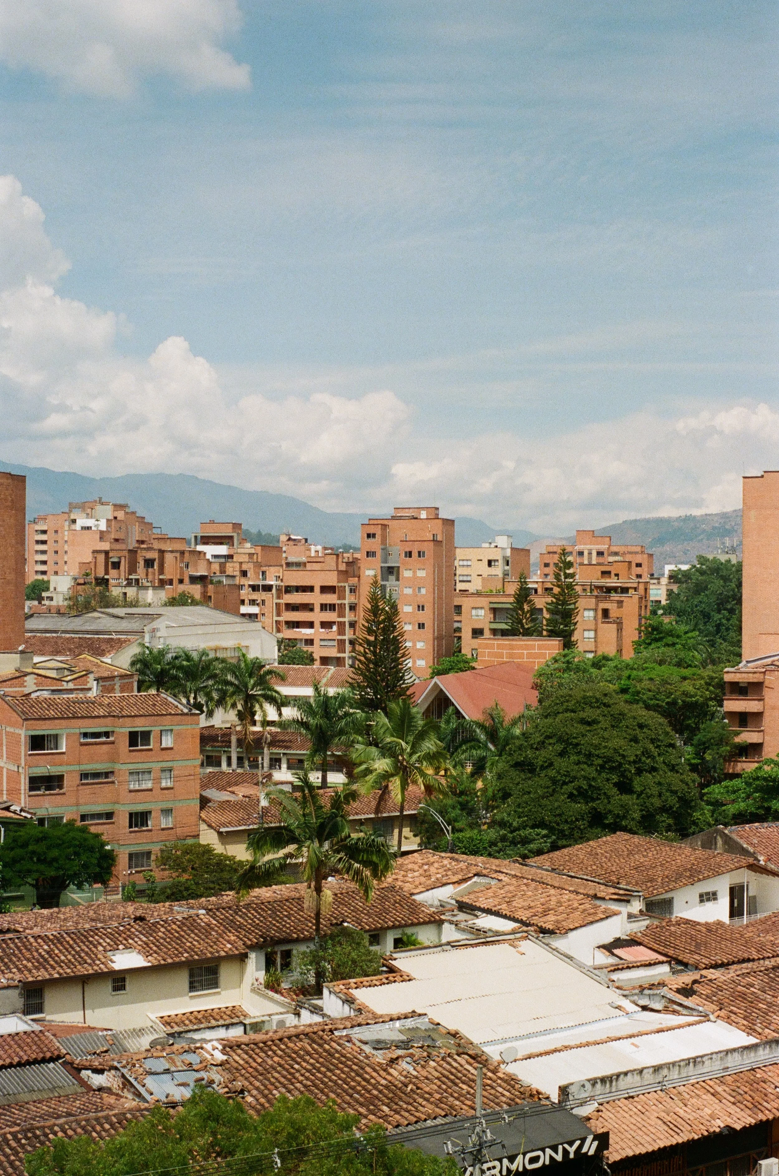 Cityscape with numerous orange-tiled rooftops, green trees, and tall apartment buildings under a partly cloudy sky.