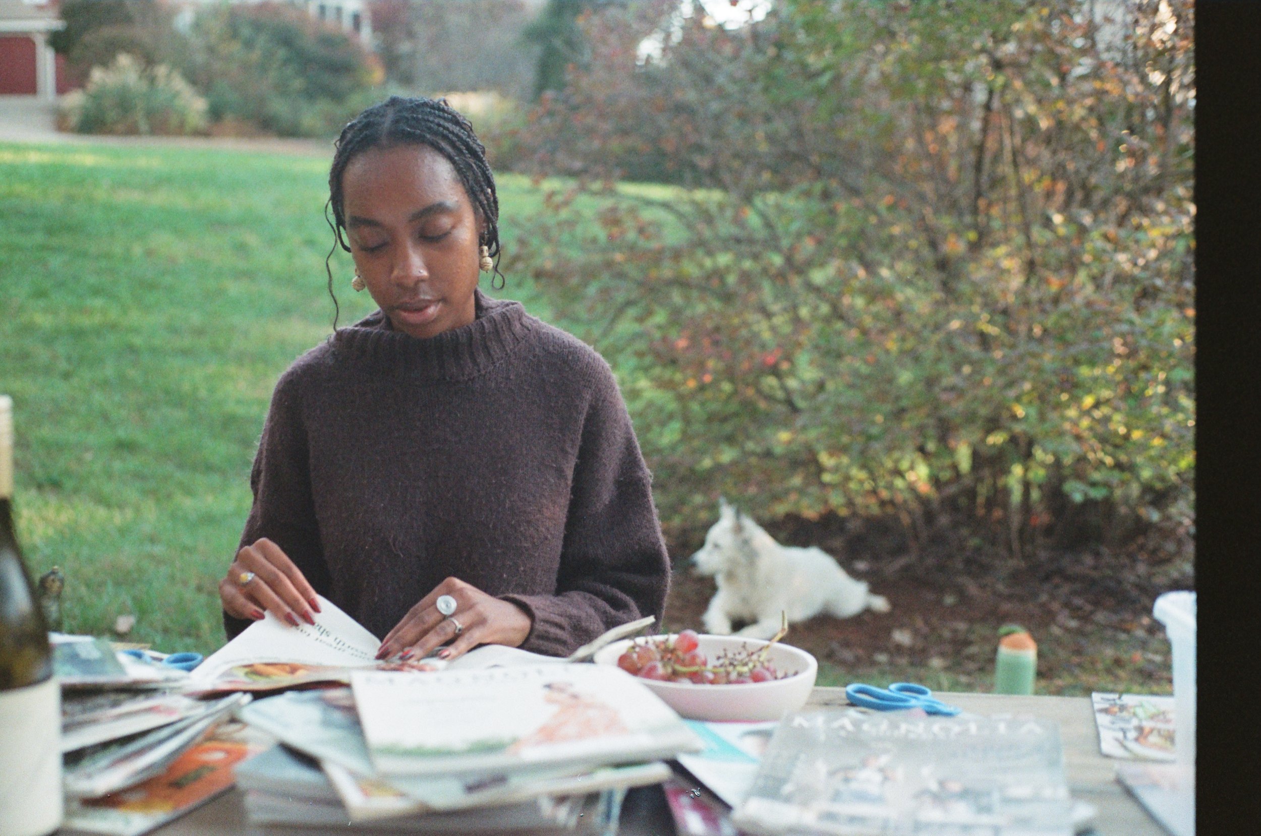 A woman with braided hair, wearing a dark brown sweater, sitting at a table outdoors, reading a magazine or catalog, with a bowl of grapes and some scattered papers and scissors on the table. A white dog lies in the background near some bushes and tr
