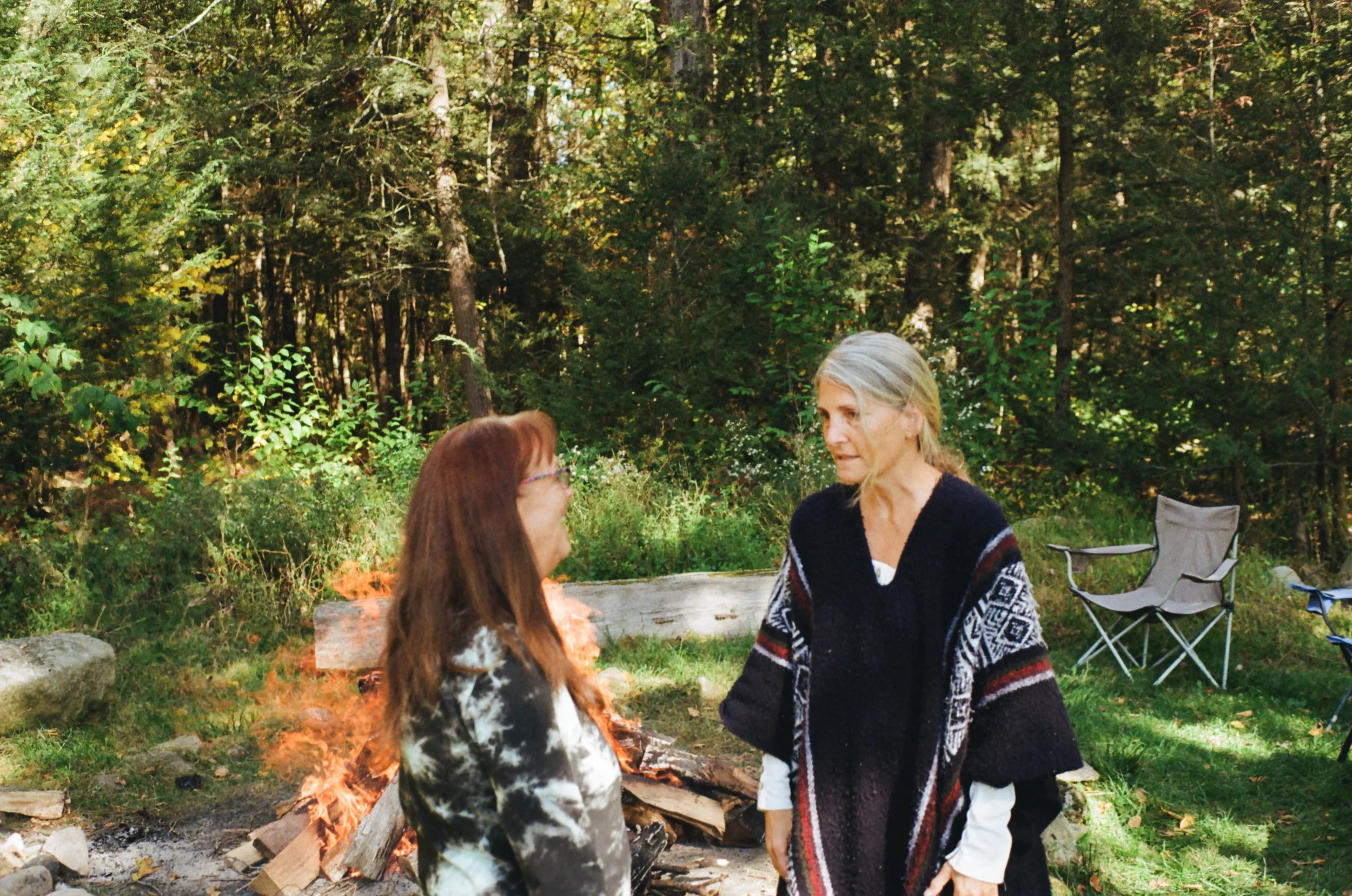 Two women talking outdoors near a campfire in a forested area with camping chairs nearby.