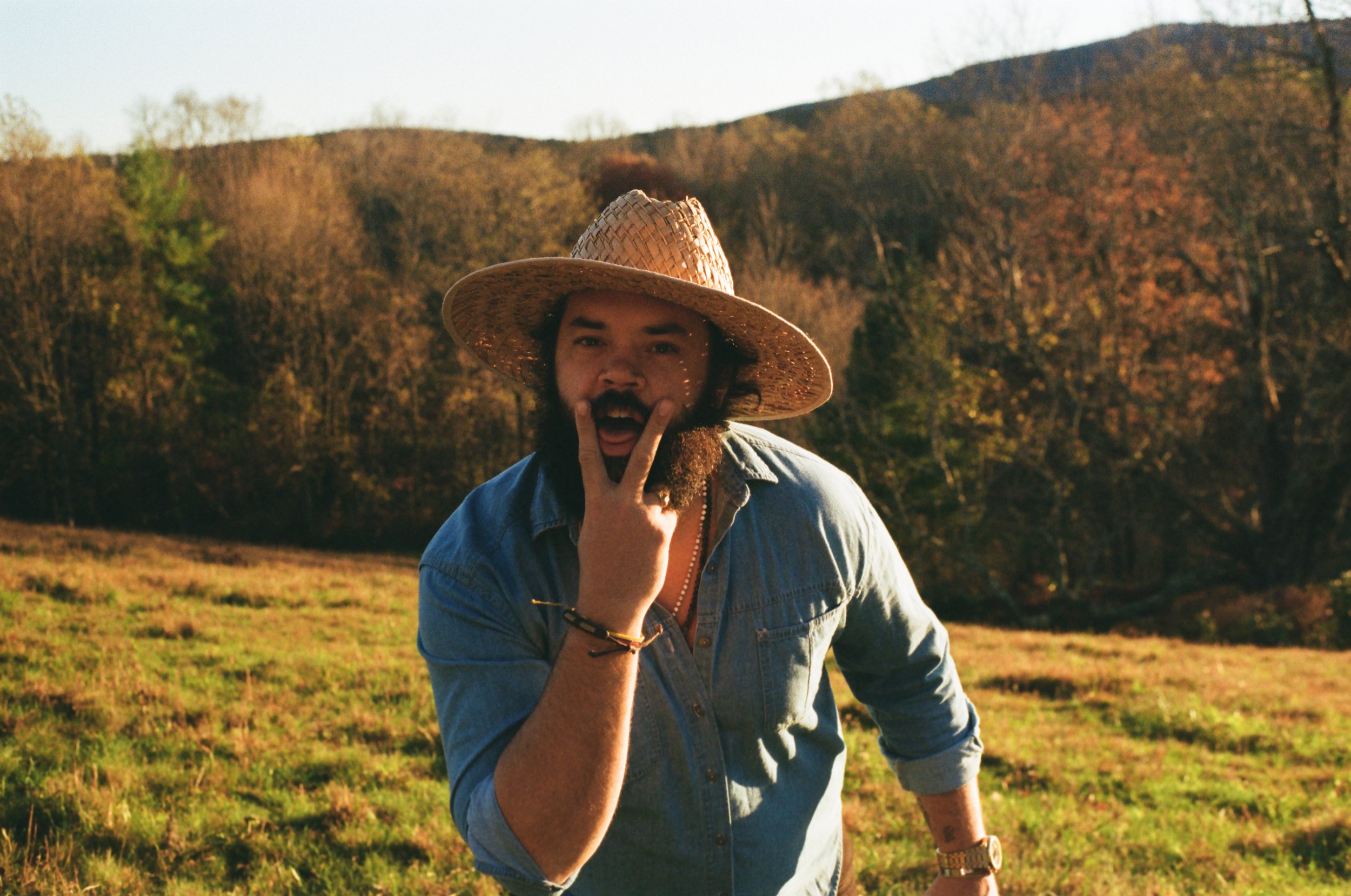 Man with beard in jean jacket and straw hat making a gesture with fingers near his face outdoors in a grassy field with trees and hills in the background