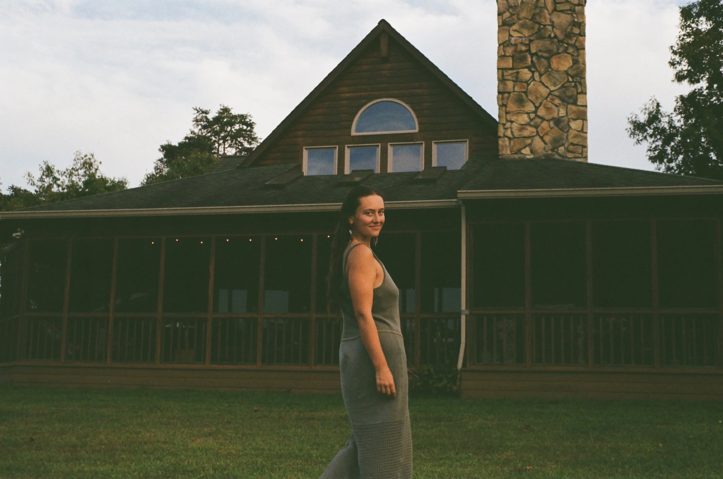 Woman in gray dress standing outside in front of a house with a stone chimney and a screened porch, trees in the background, during dusk or early evening.